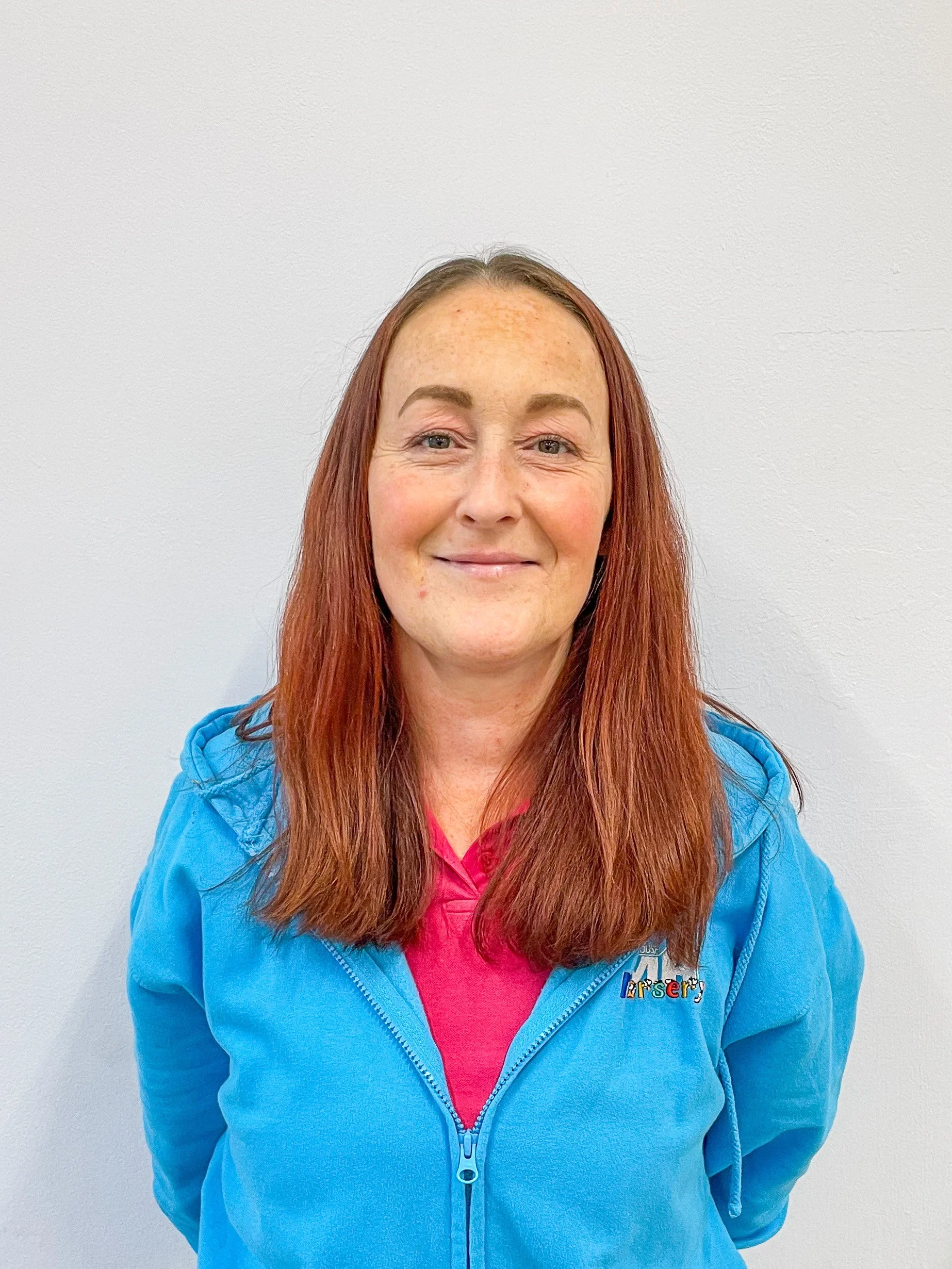 A woman with red hair and freckles, smiling, wearing a blue zip-up hoodie with a pink shirt underneath, standing against a plain white wall.
