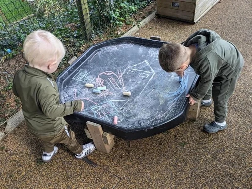 Two young boys drawing with chalk on a black tray table outdoors, surrounded by a fence and greenery.