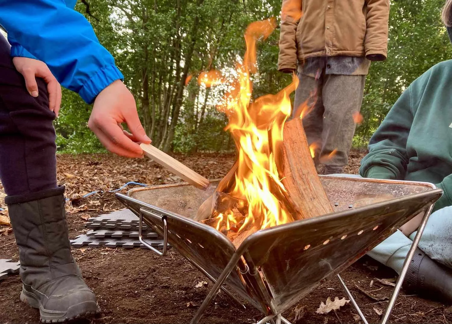 Children around a campfire in the woods, one person tending the fire with a wooden stick.