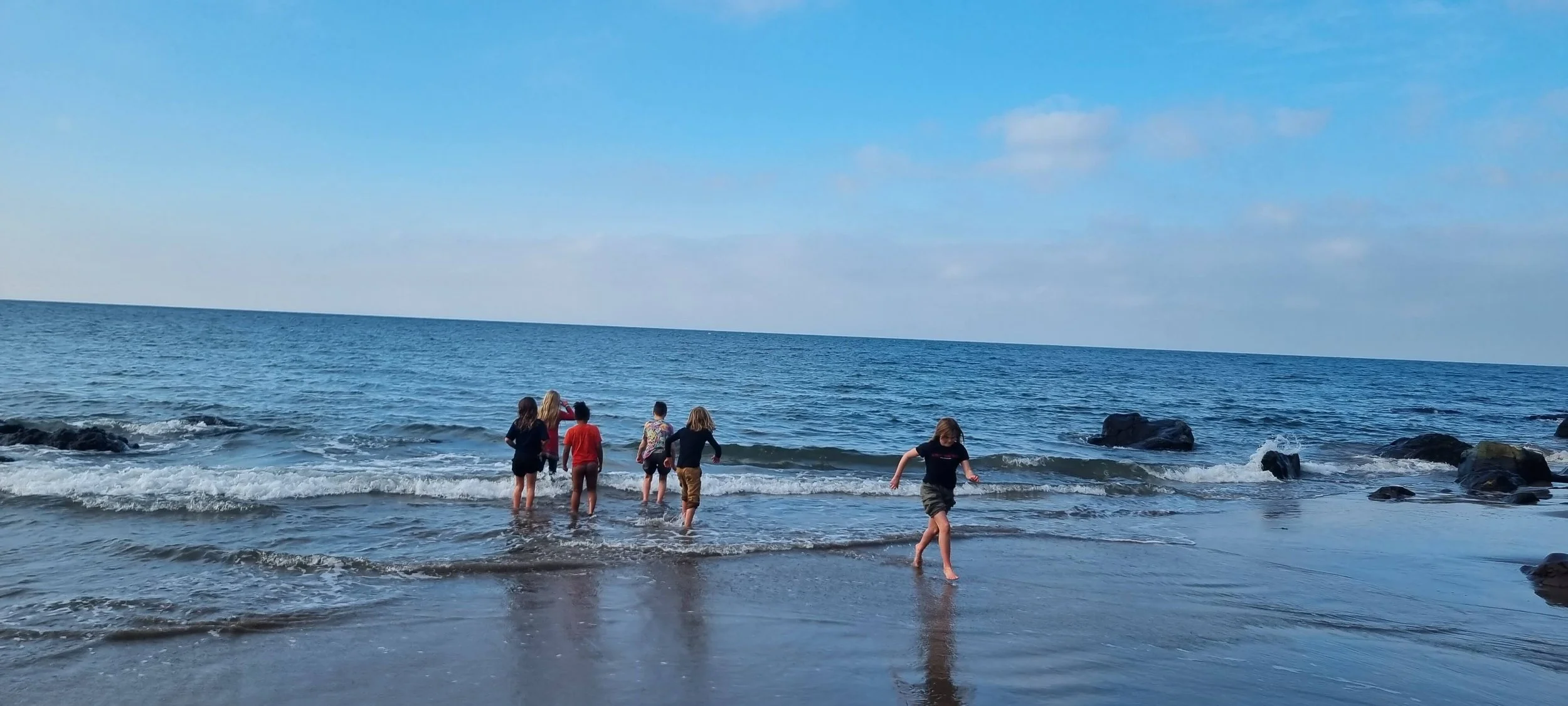 Six children playing at the shoreline of a calm ocean, with some rocks in the water, under a partly cloudy sky.