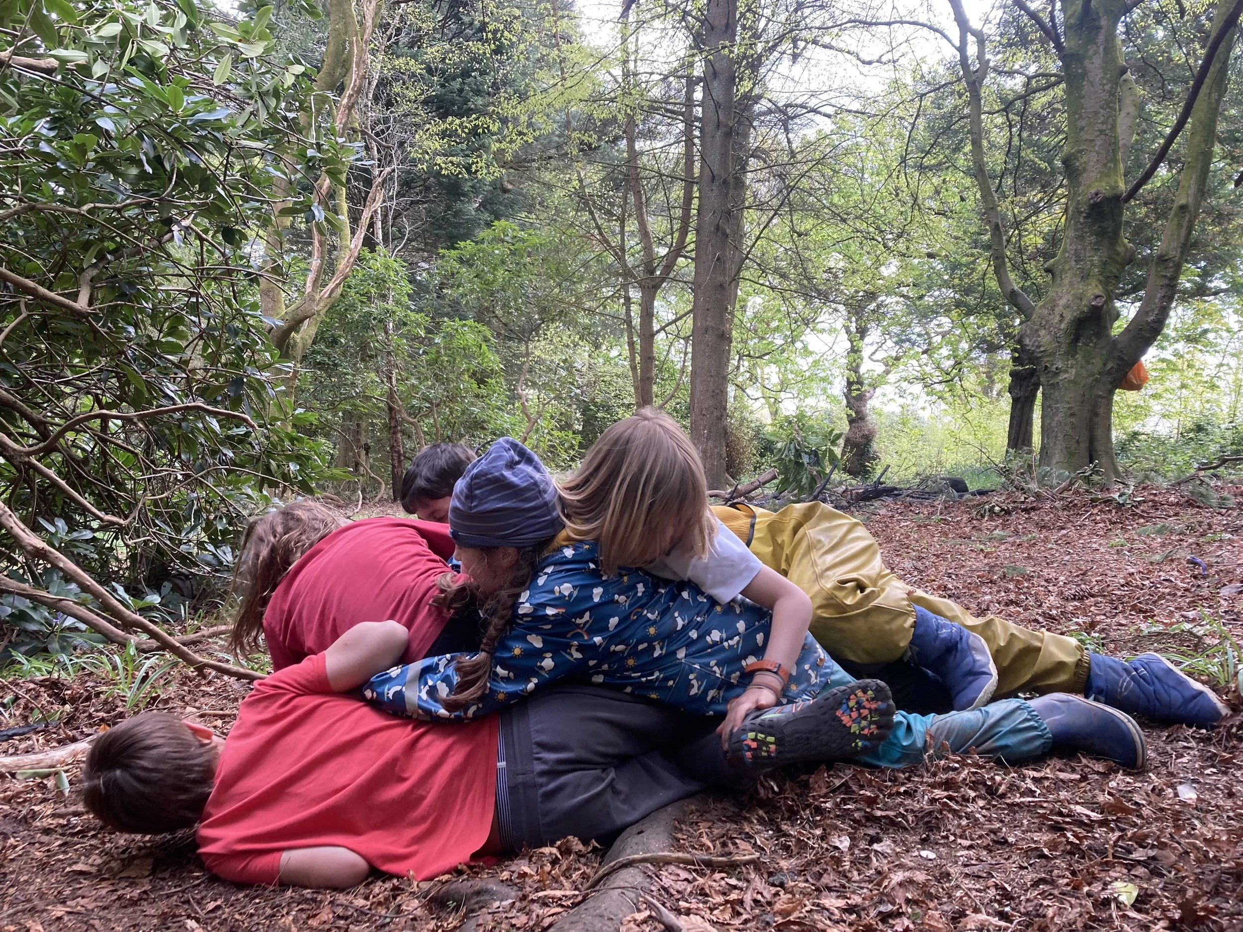 A group of children and an adult are lying on the forest ground in a pile, surrounded by trees and green foliage.