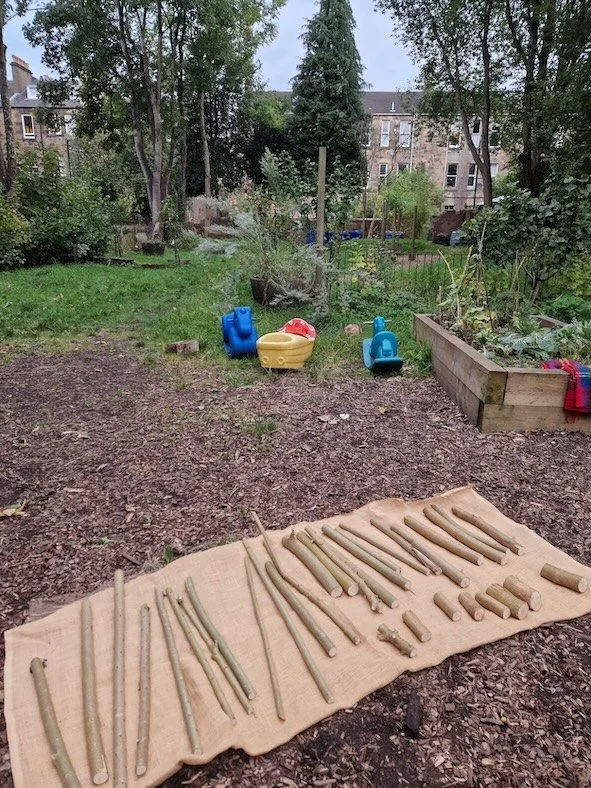 A community garden with outdoor tools laid out on a cloth, and children's watering cans and a sandbox in the background.