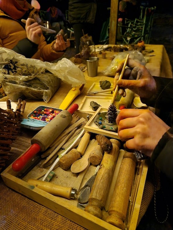 Hands working on a craft project with various tools, pinecones, and colorful beads on a wooden table.