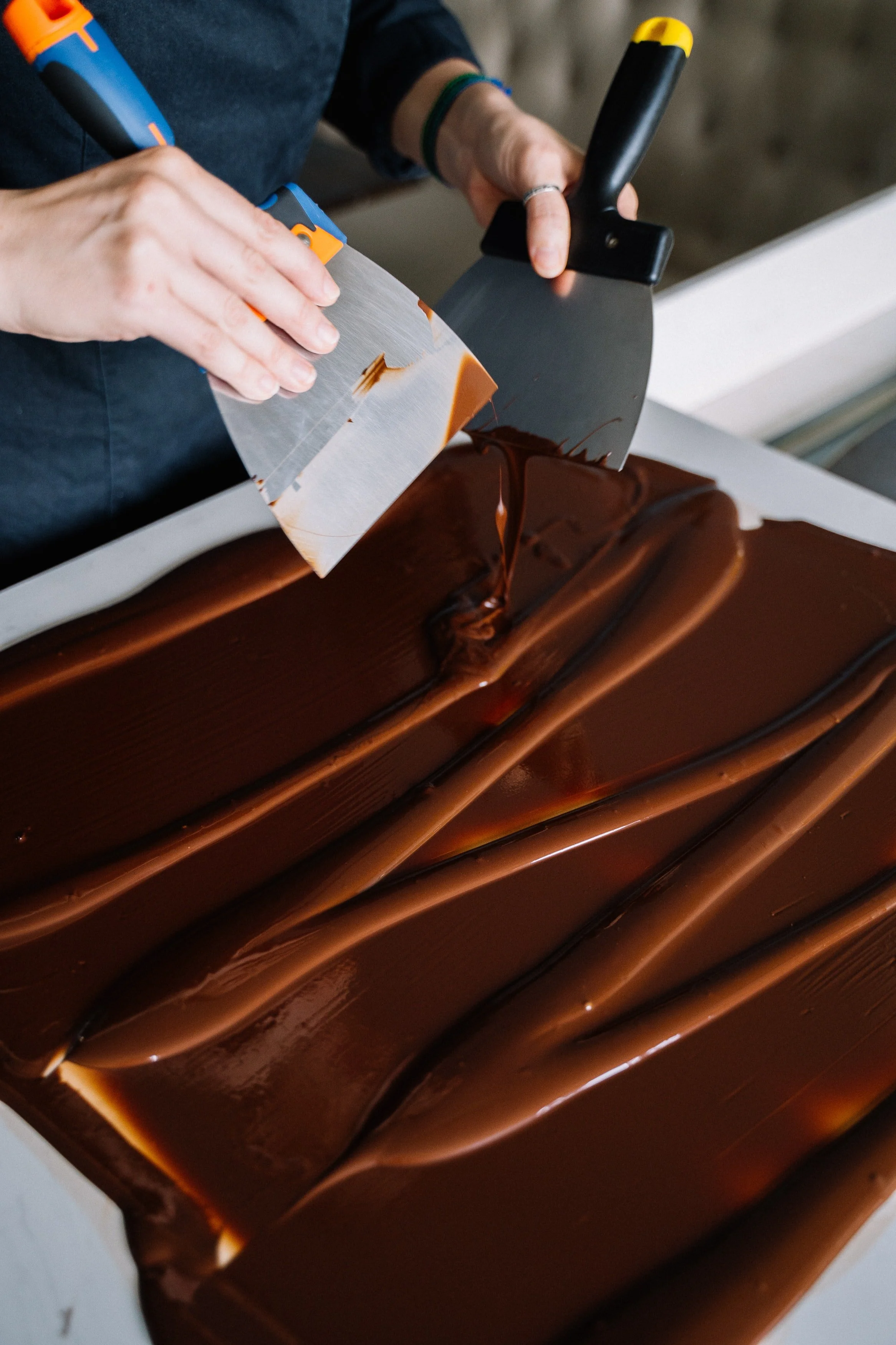 Person tempering chocolate with metal scrapers on a flat surface.