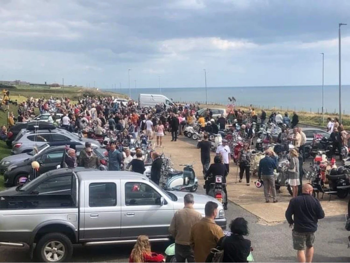 A large crowd gathered near parked motorcycles and cars by the seaside, with people socializing and admiring the vehicles.