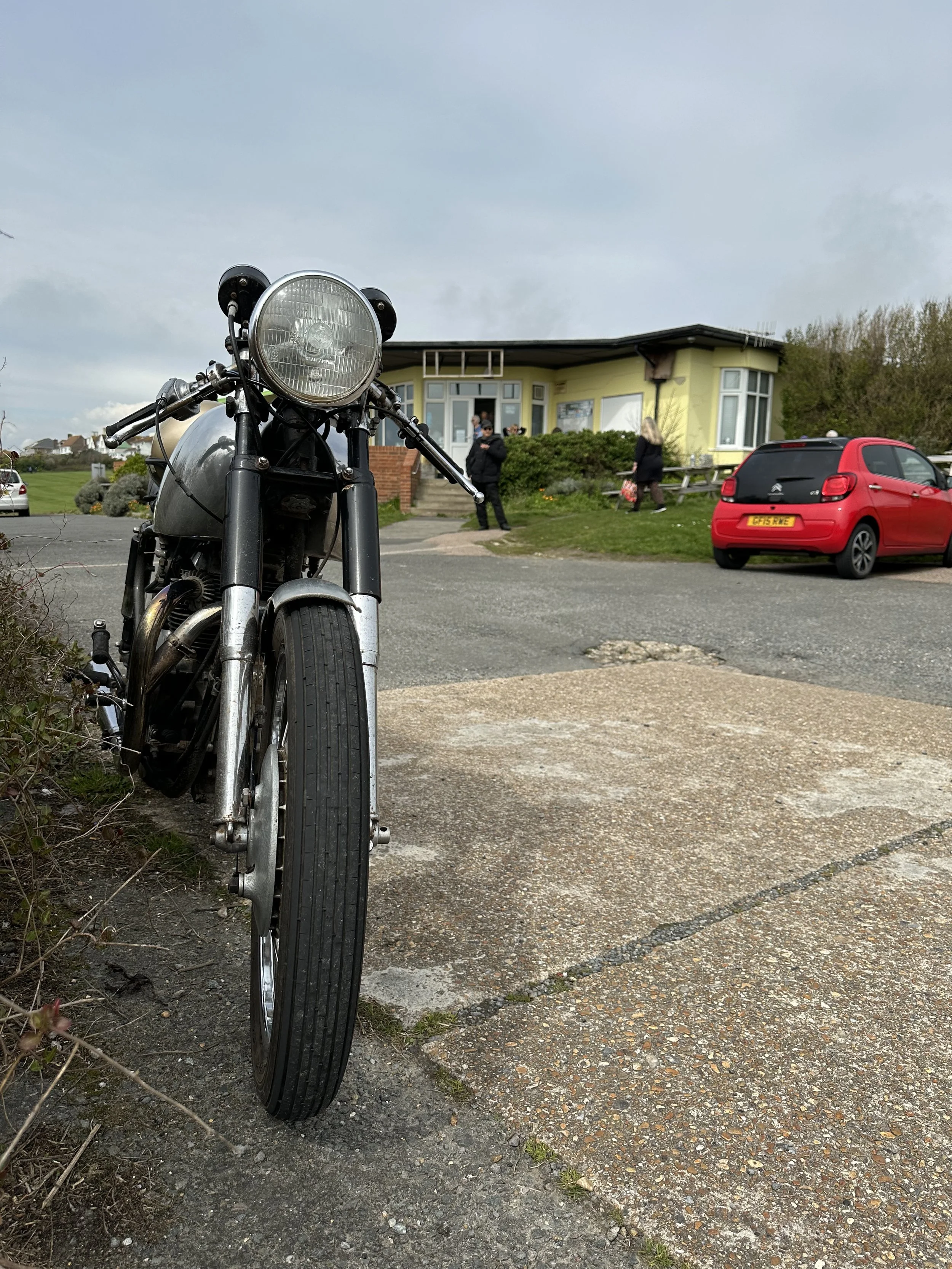 A vintage motorcycle parked on a driveway, with a yellow house and parked red car in the background. 