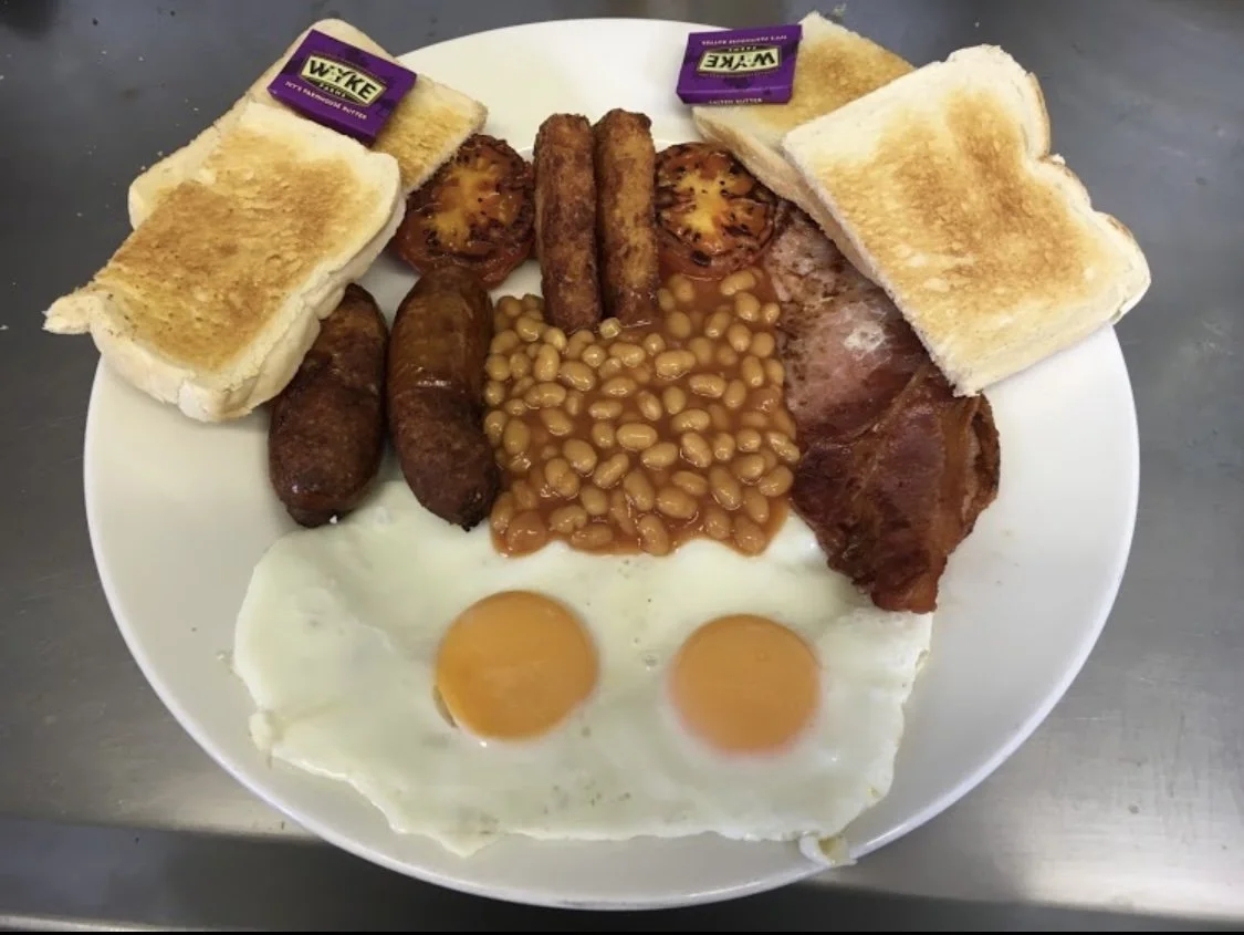 English breakfast with eggs, sausages, bacon, baked beans, hash browns, grilled tomato, toast, and butter packets on a white plate.