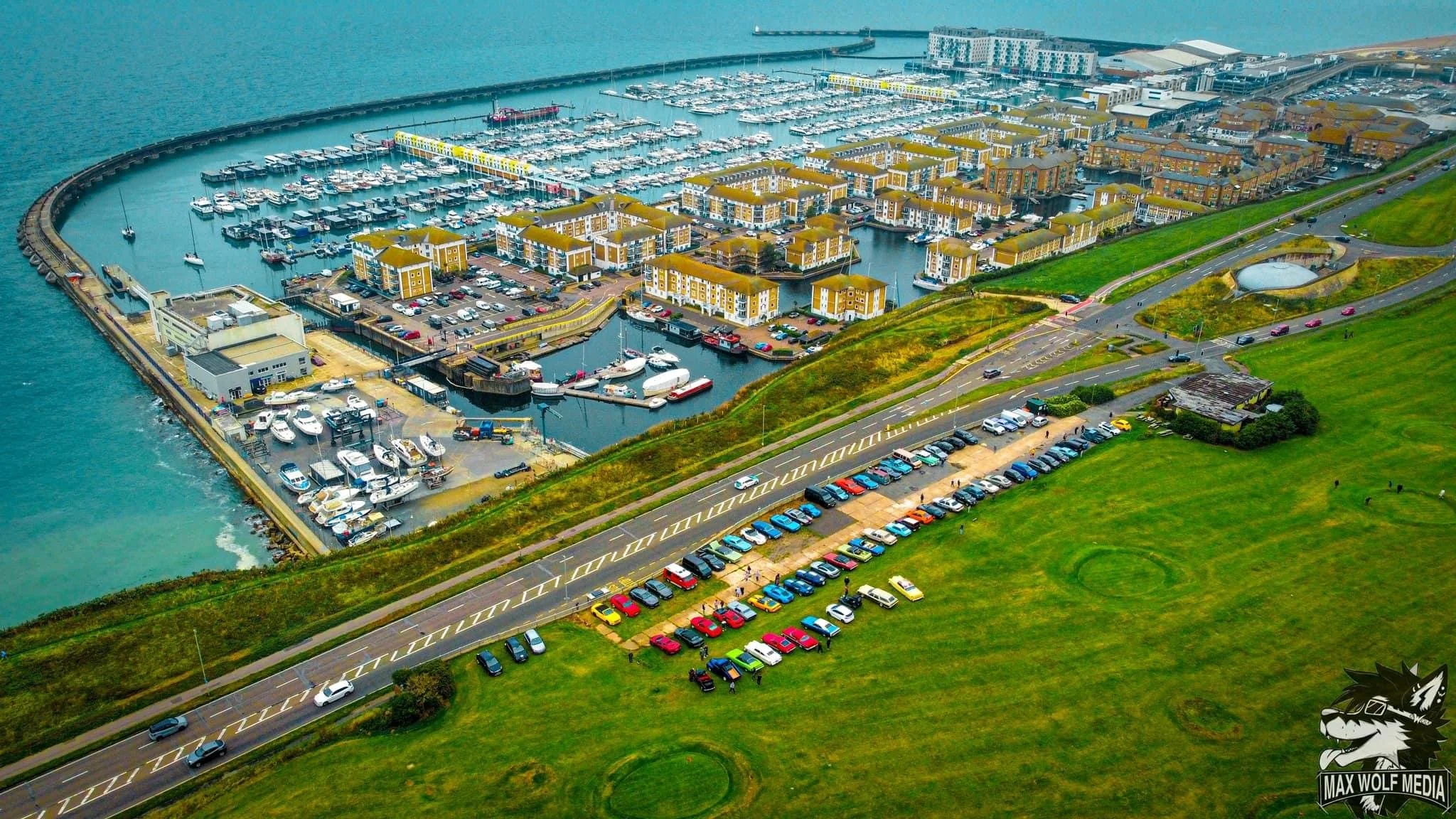Aerial view of a marina with boats, surrounding residential buildings, parking lot, and grassy area. A coastal road runs through the landscape near the sea.