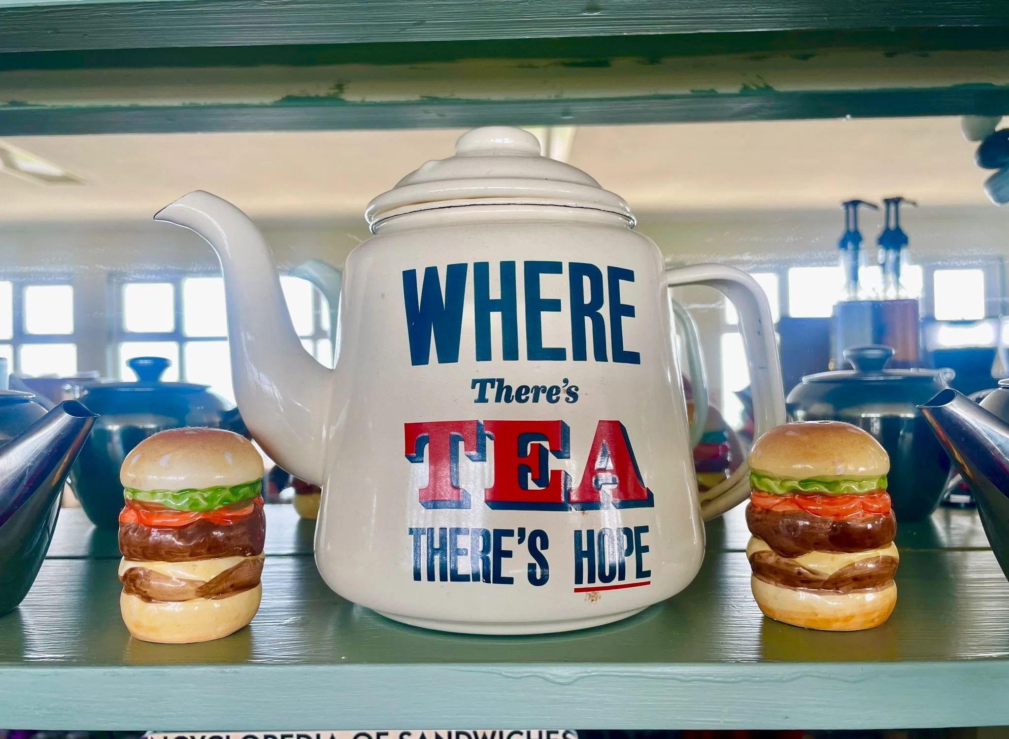 Teapot with quote "Where there's tea there's hope" next to two ceramic burger figurines on a shelf.