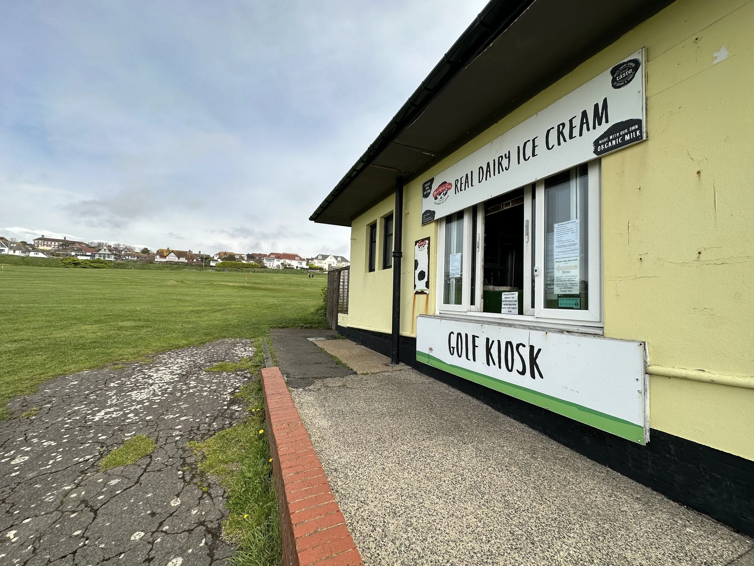 Exterior of an ice cream and golf kiosk with signs for real dairy ice cream, showing an open window. A grassy lawn and houses in the distance under a cloudy sky.