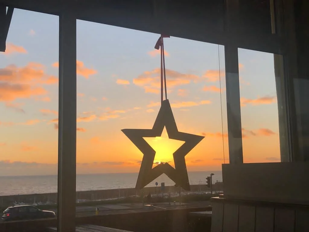 A star-shaped decoration hanging in a window with a sunset and ocean view in the background.