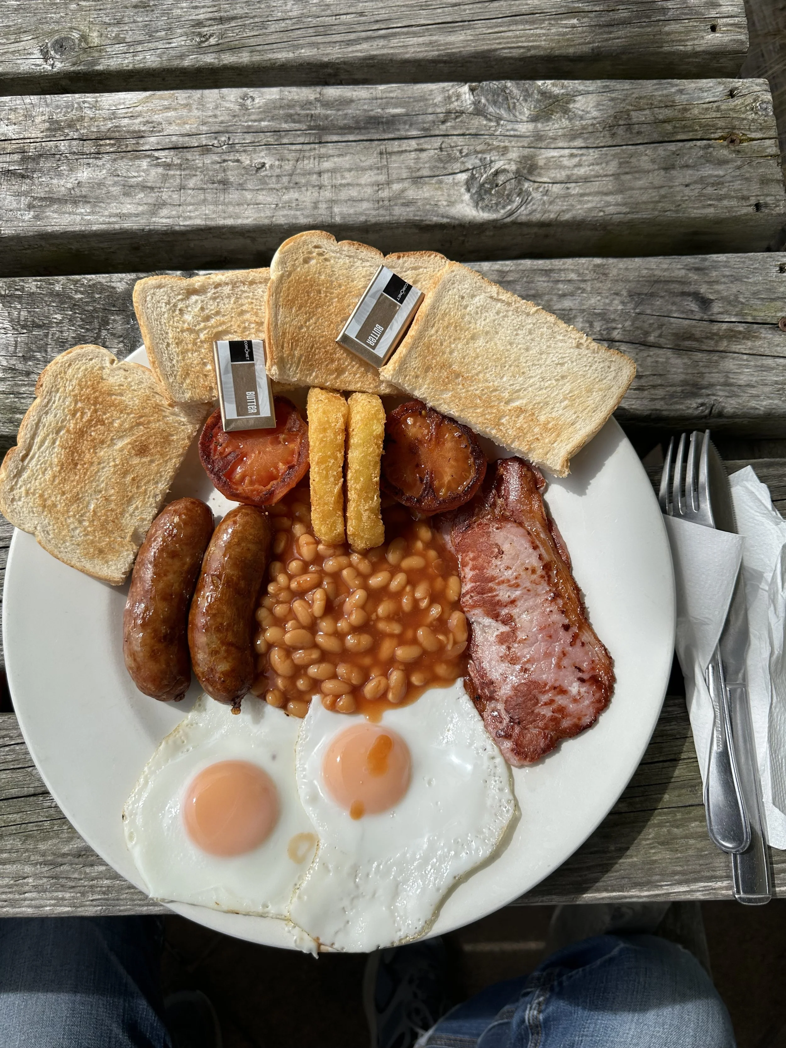Plate of a full English breakfast with fried eggs, sausages, bacon, baked beans, grilled tomato, hash browns, and slices of toast with butter packets, on a wooden table.
