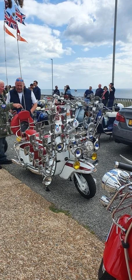 A group of  Mods with vintage scooters at our Brighton seafront location. The scooters are adorned with multiple mirrors and headlights, as is the fashion.. We host regular scooter, bike and classic car meets