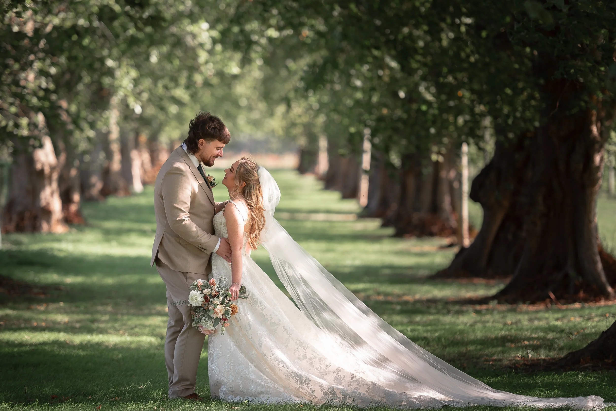 A bride and groom standing close together outdoors, holding hands and looking at each other affectionately, with green trees in the background.
