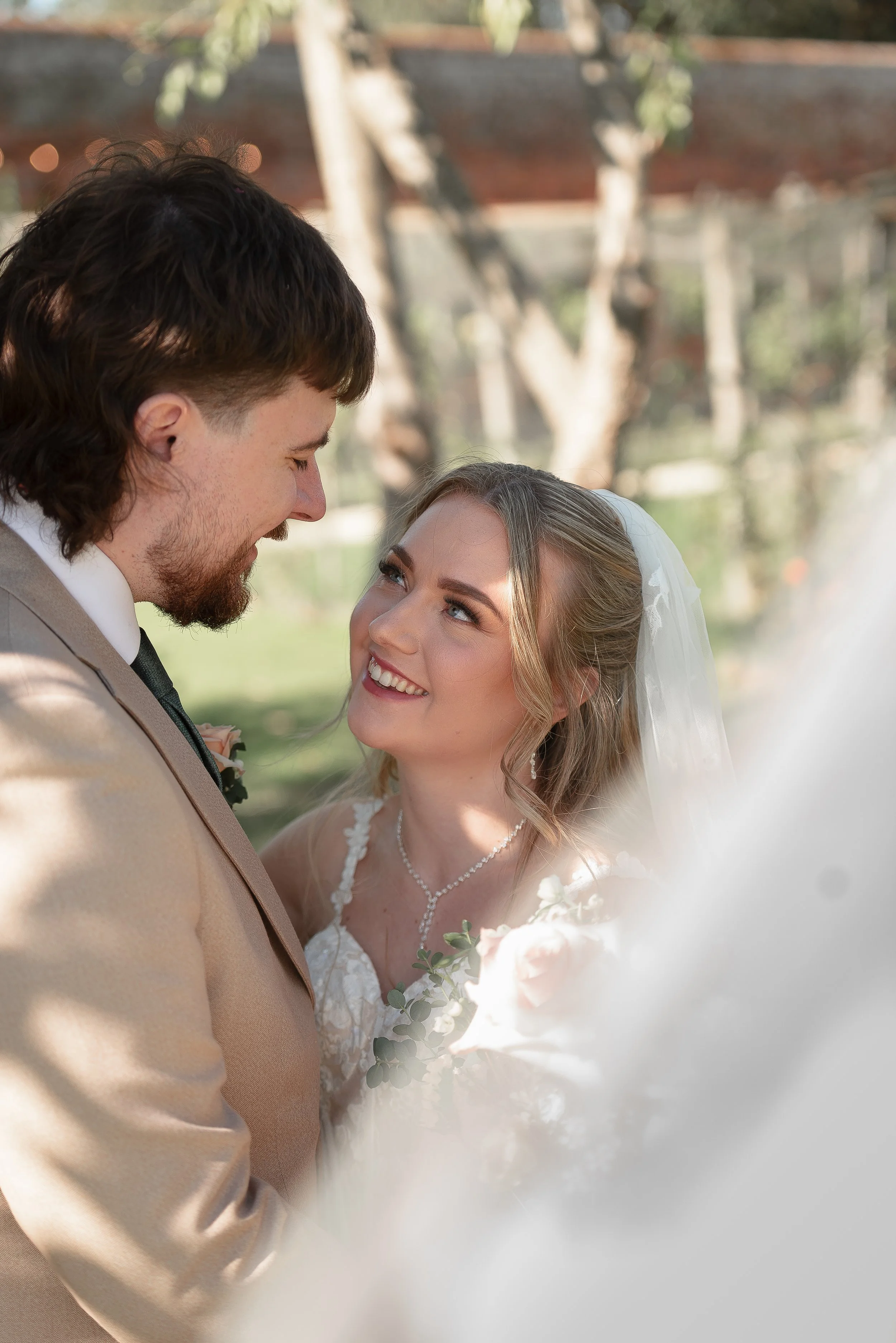 A bride and groom are looking at each other smiling outdoors, with blurred trees and a building in the background, during their wedding ceremony.