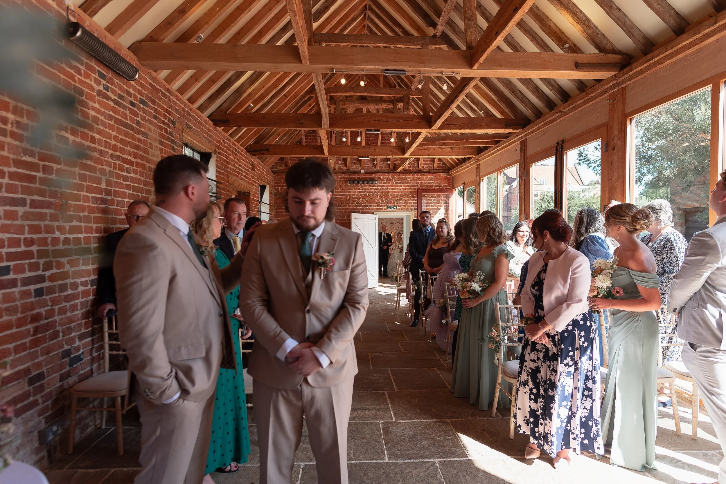 People dressed in formal attire attending a wedding ceremony inside a rustic brick and wood venue with large windows.