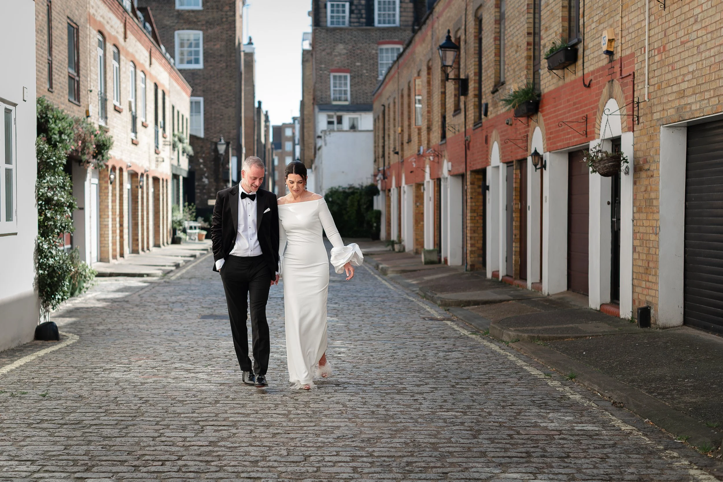 A bride and groom in wedding attire walking arm in arm down a cobblestone street lined with brick buildings outside Marylebone Town Hall after getting married.