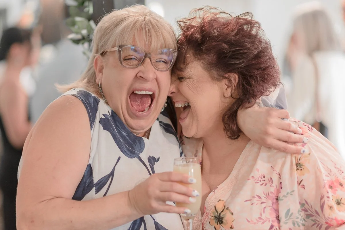 Mother of the bride and mother of the groom are laughing and embracing at the wedding breakfast, with one holding a glass of white wine.