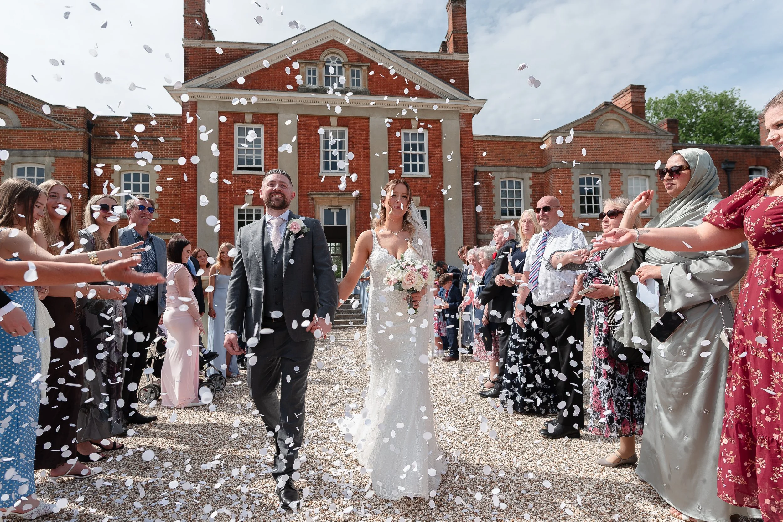 A bride and groom walking out of a building after their wedding ceremony, surrounded by guests throwing confetti, with a red brick building in the background.