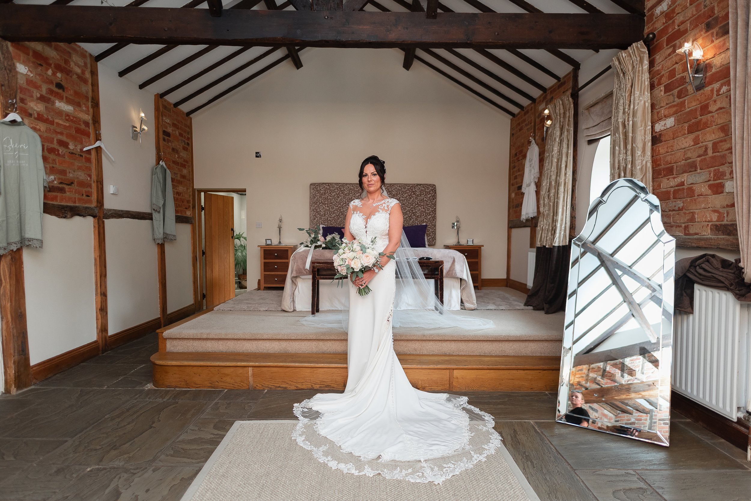 A bride in a white wedding dress holding a bouquet of flowers, standing in a rustic interior room with exposed brick walls and a high ceiling with dark wooden beams, in front of a bed with purple pillows and a padded headboard.
