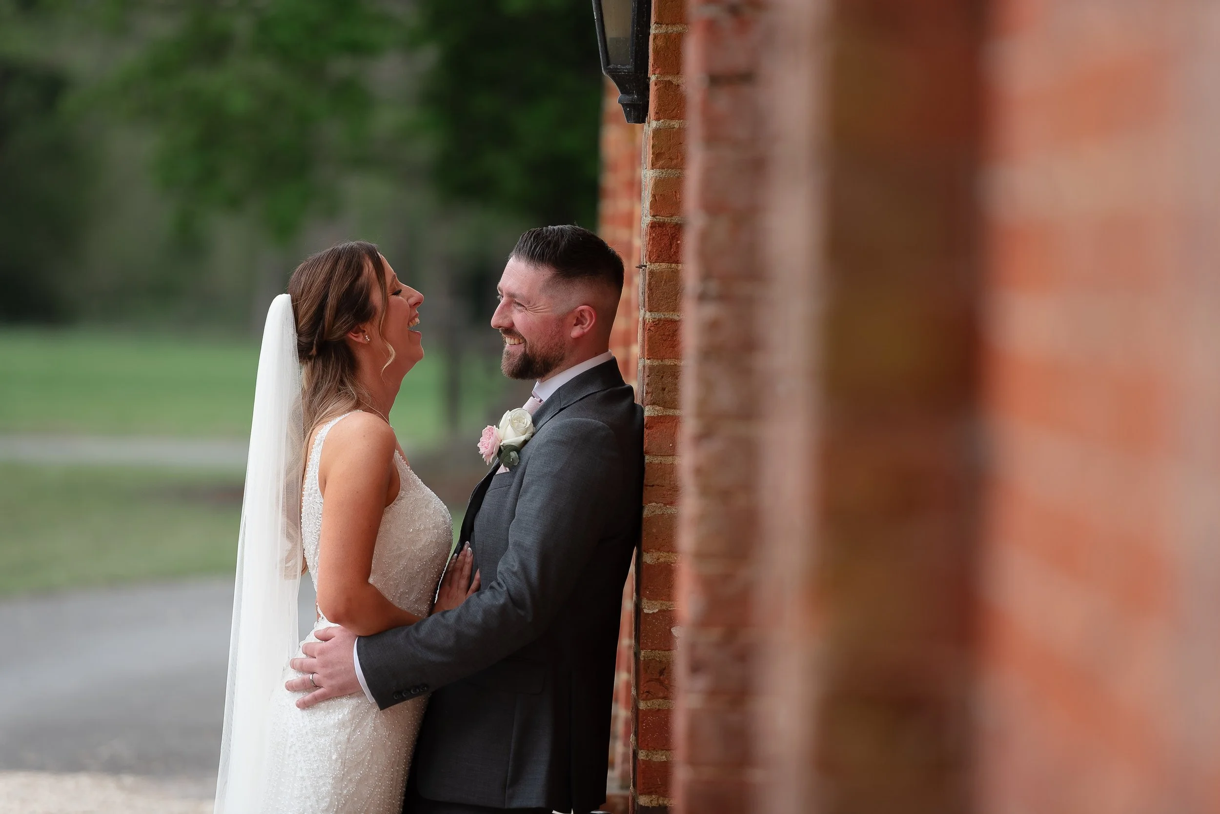 A bride and groom smiling and looking at each other, standing close to a brick wall outdoors during a wedding.
