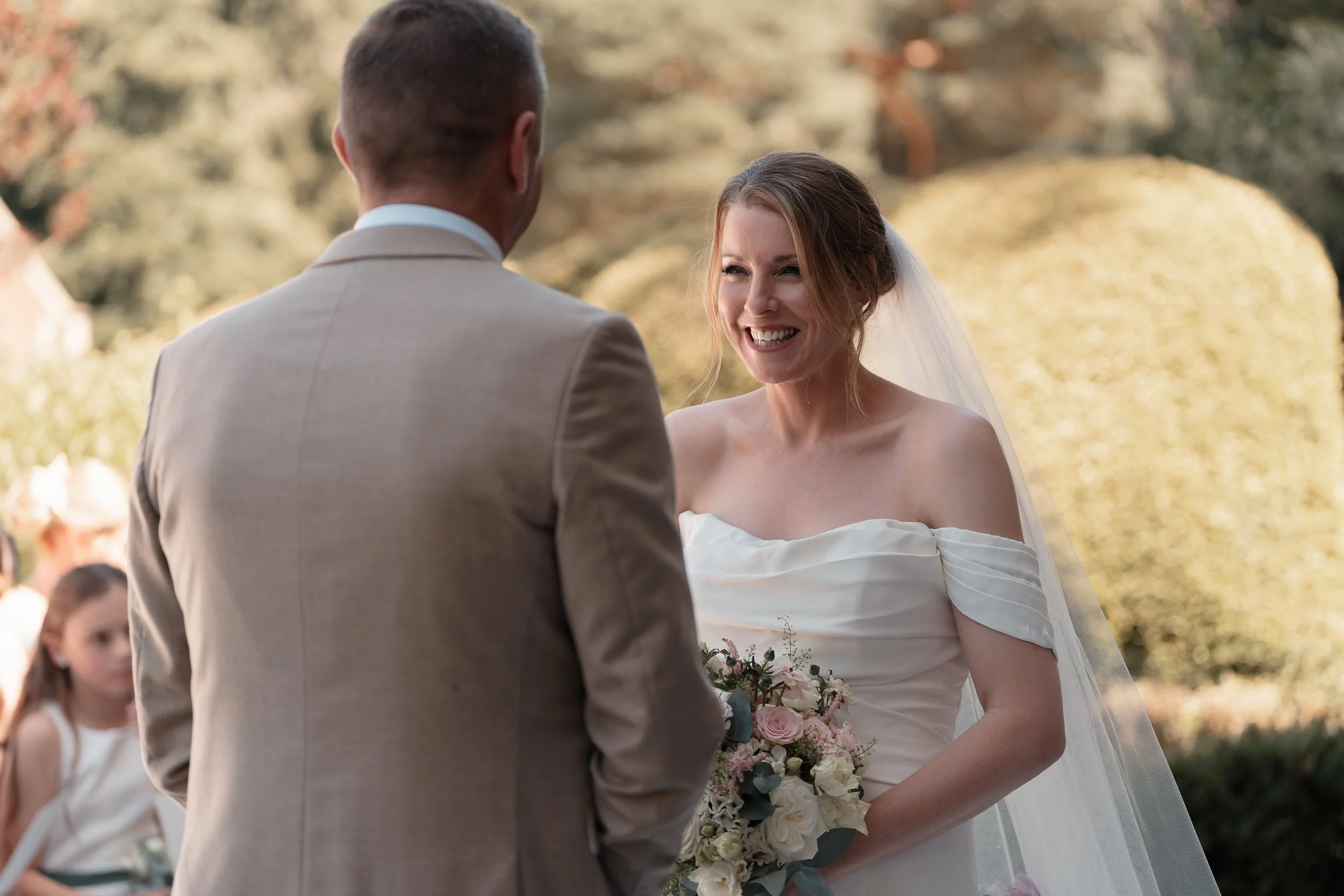 A bride and groom exchanging vows outdoors during a wedding ceremony, with guests seated in the background.