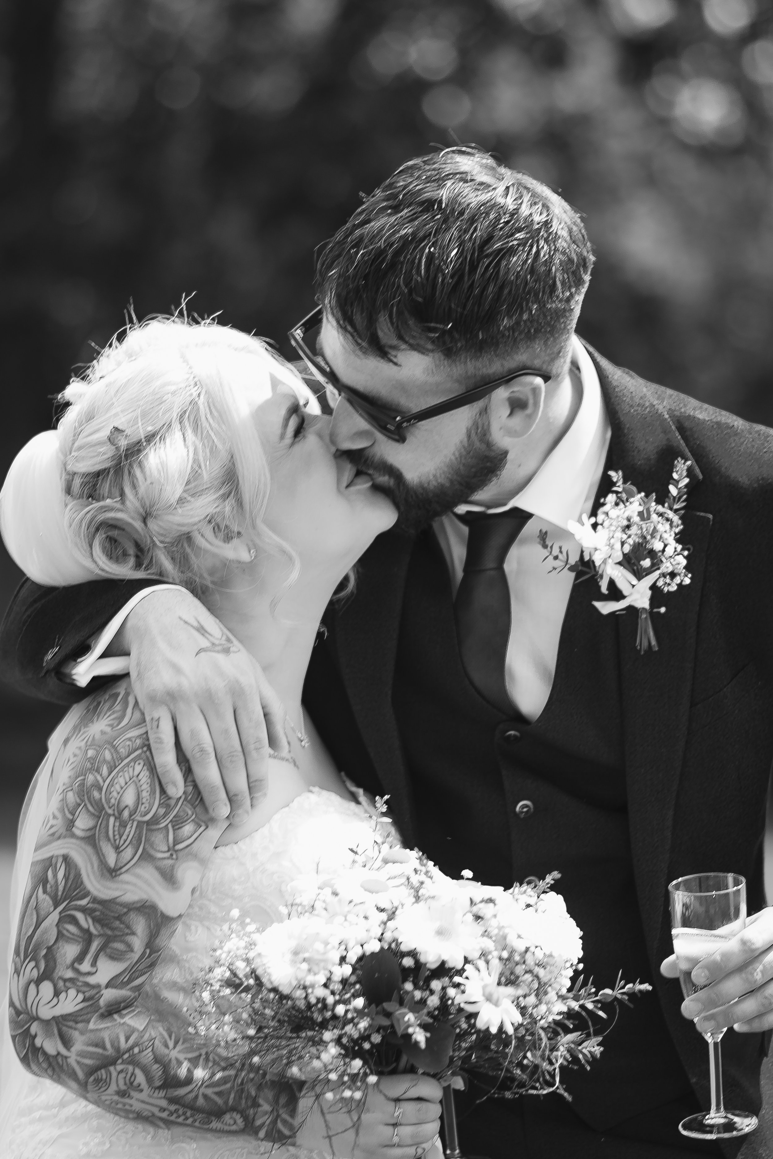 A newlywed couple sharing a kiss at their wedding reception. The bride has platinum blonde hair and tattoos on her arm. The groom is wearing glasses and a boutonniere on his tuxedo. There is a floral arrangement and a toast glass in the foreground.