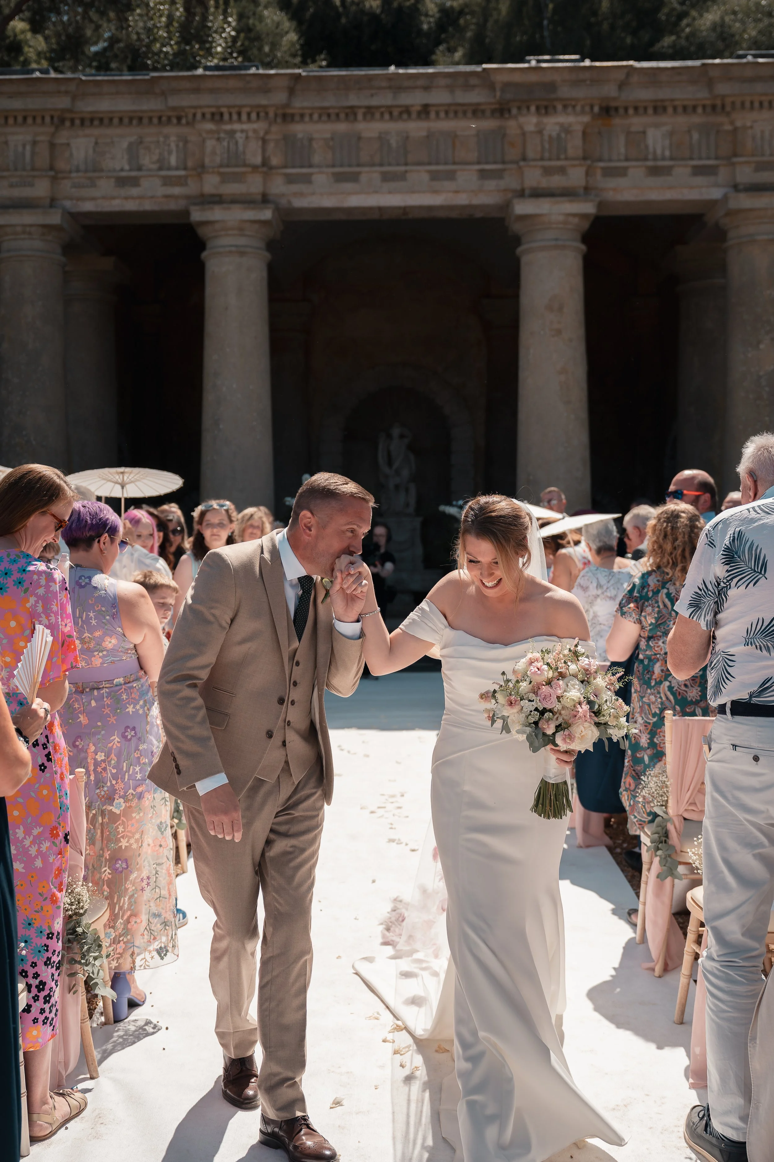 A bride in a white wedding dress holding a bouquet walking down the aisle of an outdoor wedding ceremony, with groom and guests around, set against a historic stone building with columns.