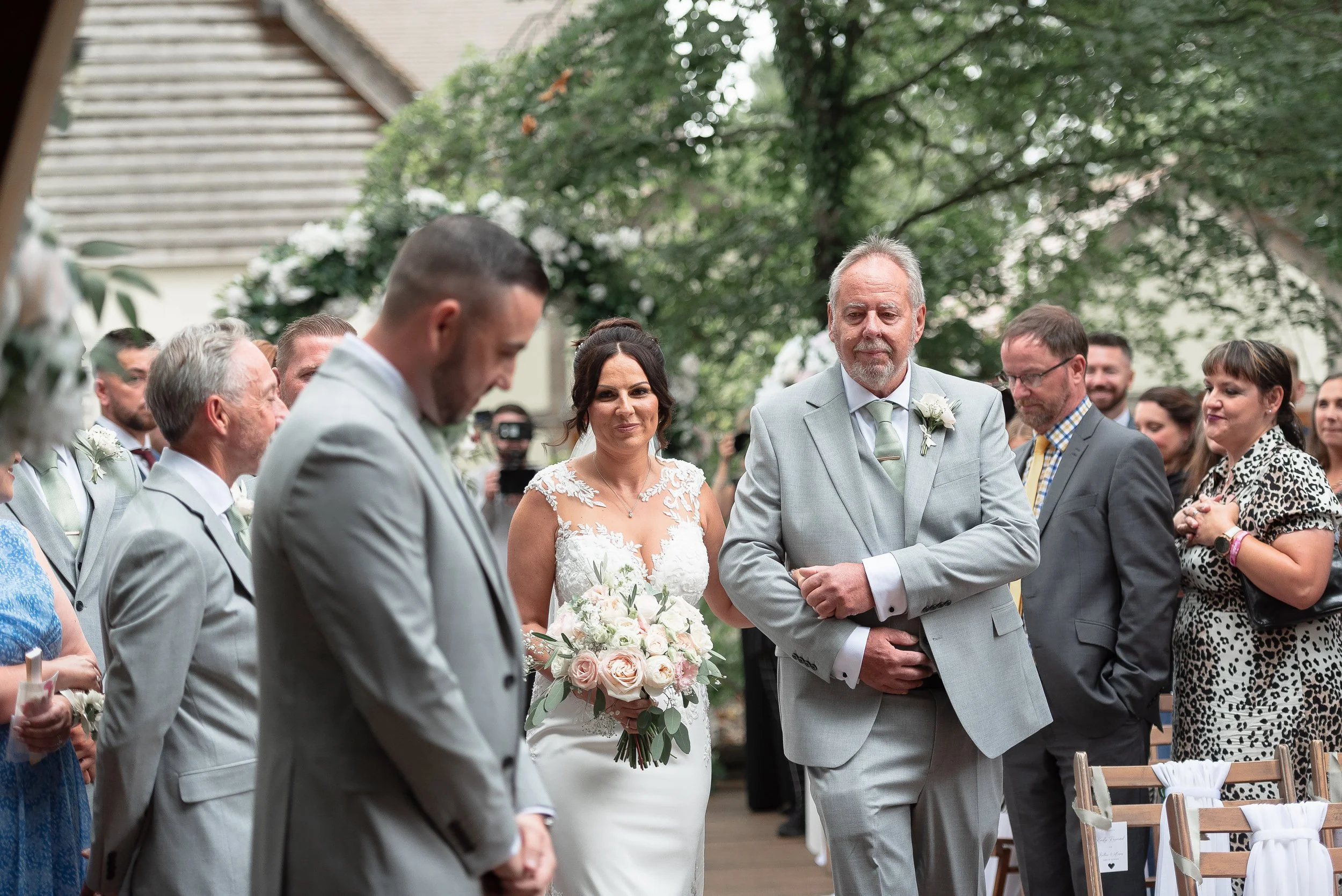 A wedding ceremony outdoors with the bride and groom standing among friends and family, surrounded by trees and greenery, with wedding chairs and flowers visible.