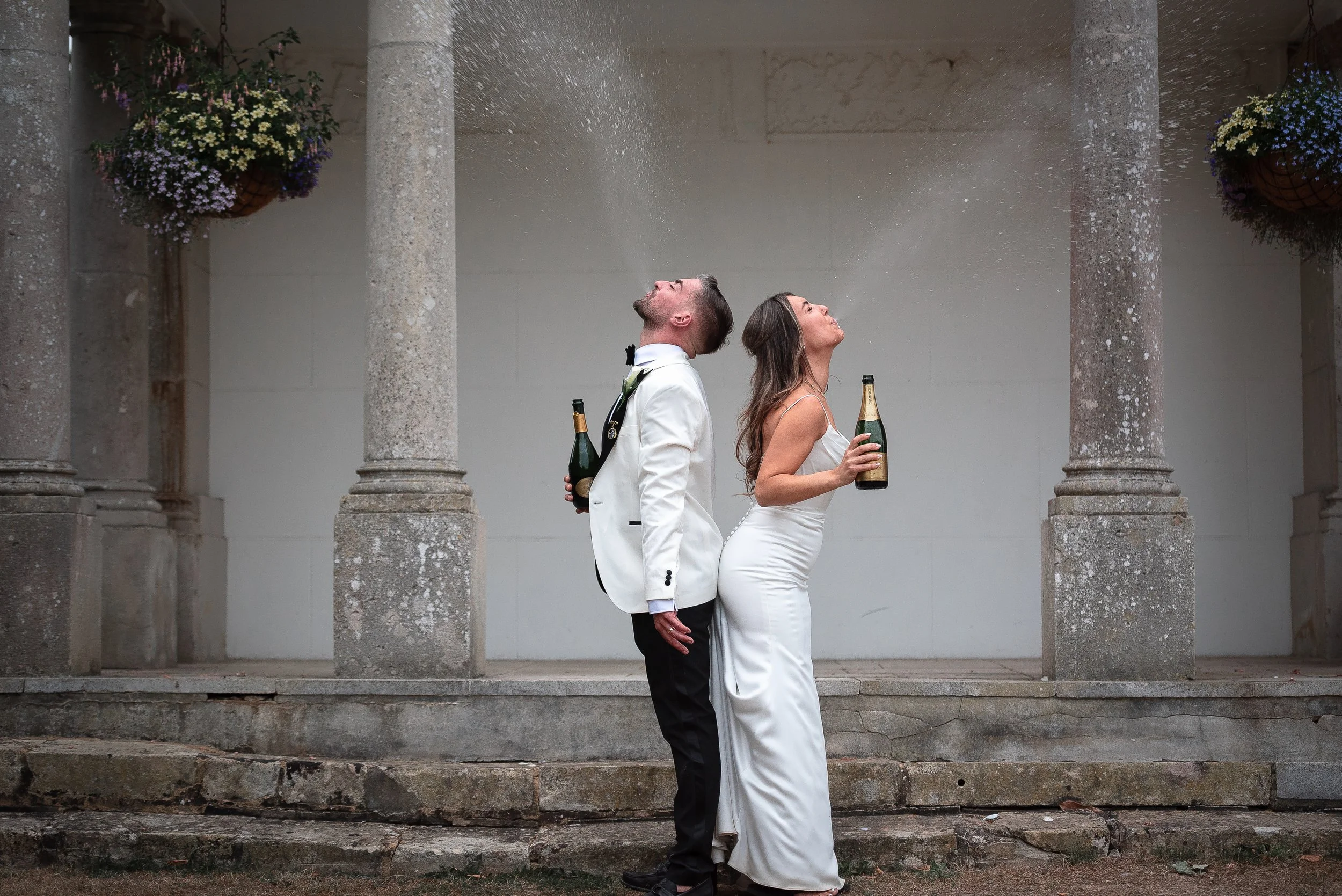 A bride and groom, dressed in wedding attire, standing back to back and holding champagne bottles, spraying champagne out of their mouths at a Hampshire wedding venue, Highfield Park