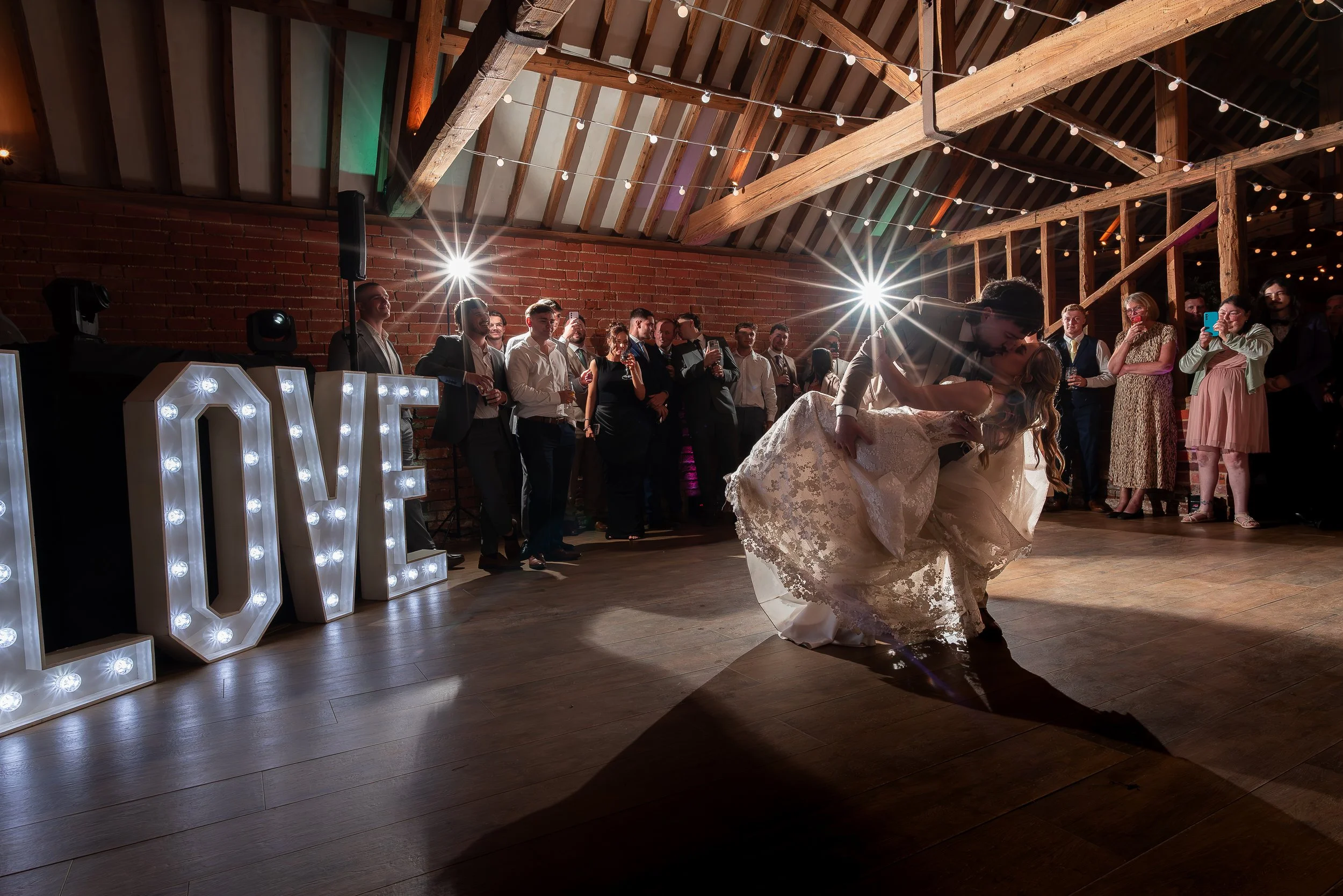 At a wedding reception, a bride and groom are dancing on the wooden floor while guests watch and take photos. The venue has a brick wall, wooden beams, string lights, and illuminated marquee letters spelling 'LOVE'.