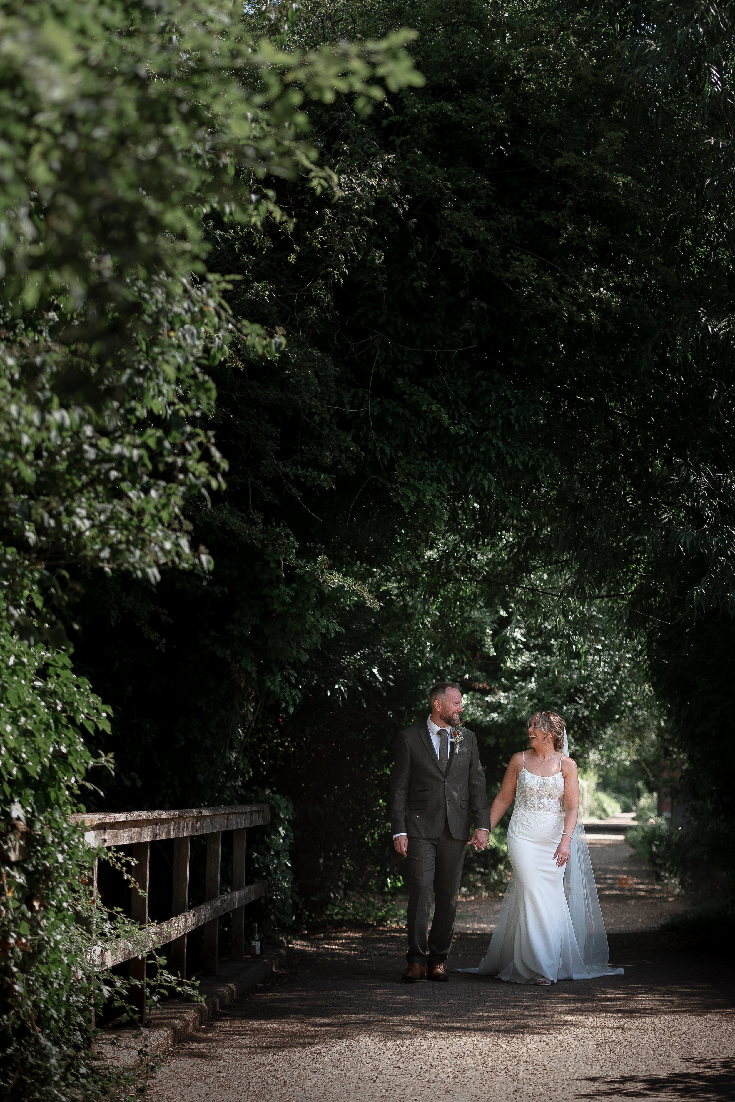 A newlywed couple walking hand in hand on a shaded path surrounded by lush green trees.