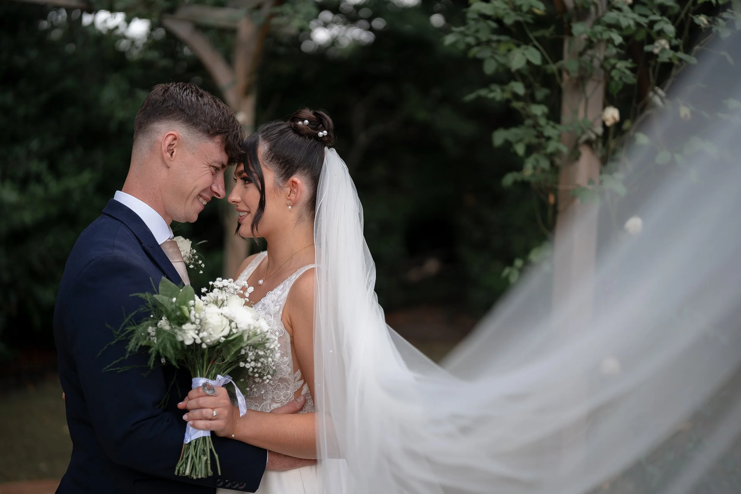 A bride and groom standing close together outdoors, smiling with foreheads touching, during their wedding ceremony. The bride has dark hair styled in an updo with pearl accessories and a white veil, and she is holding a bouquet of white flowers. The groom has short brown hair styled neatly and is wearing a navy blue suit with a light-colored tie. Green foliage and trees are visible in the background.