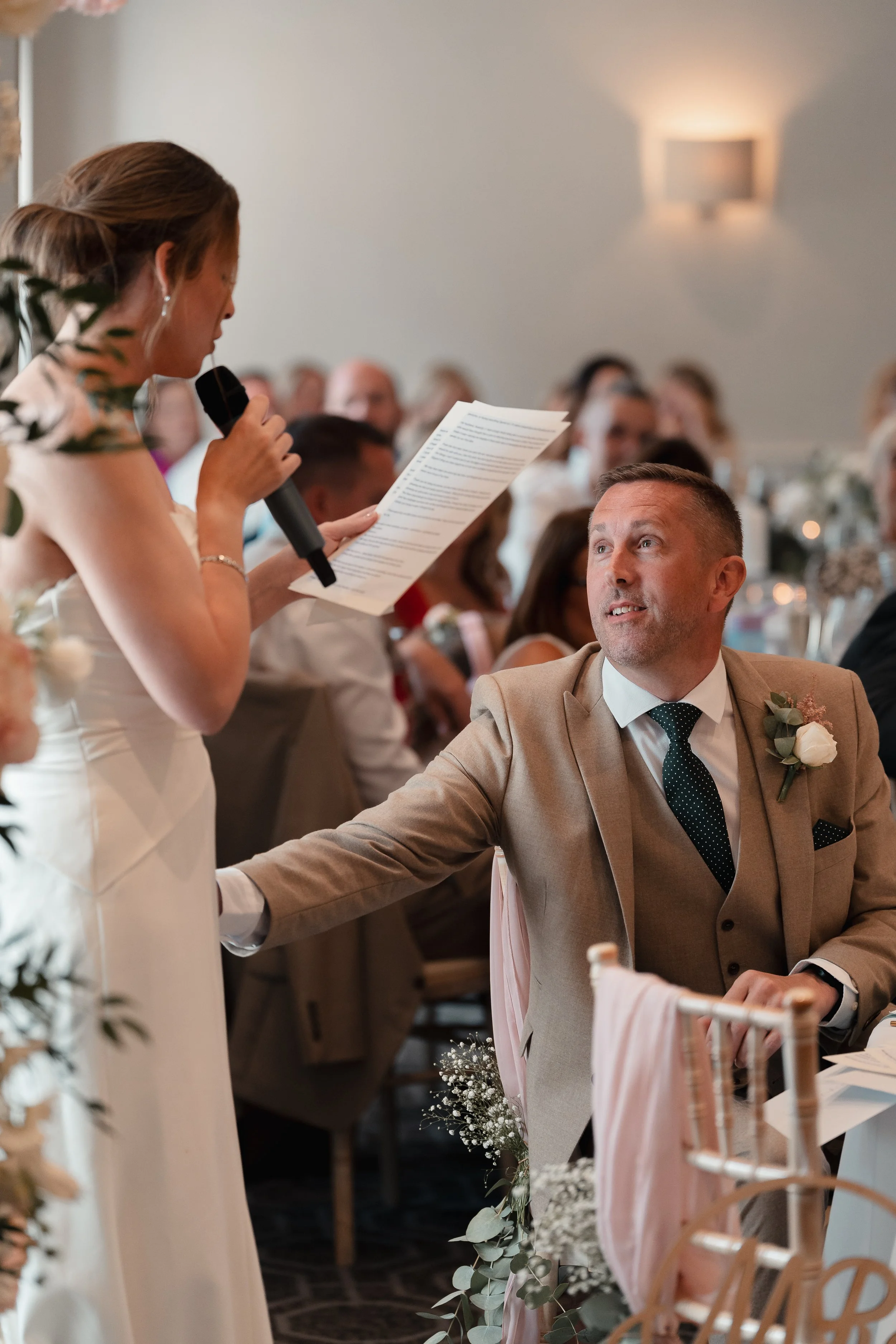A woman giving a speech at a wedding reception, holding a microphone and reading from a paper, while a man, possibly the groom, sitting and listening attentively.