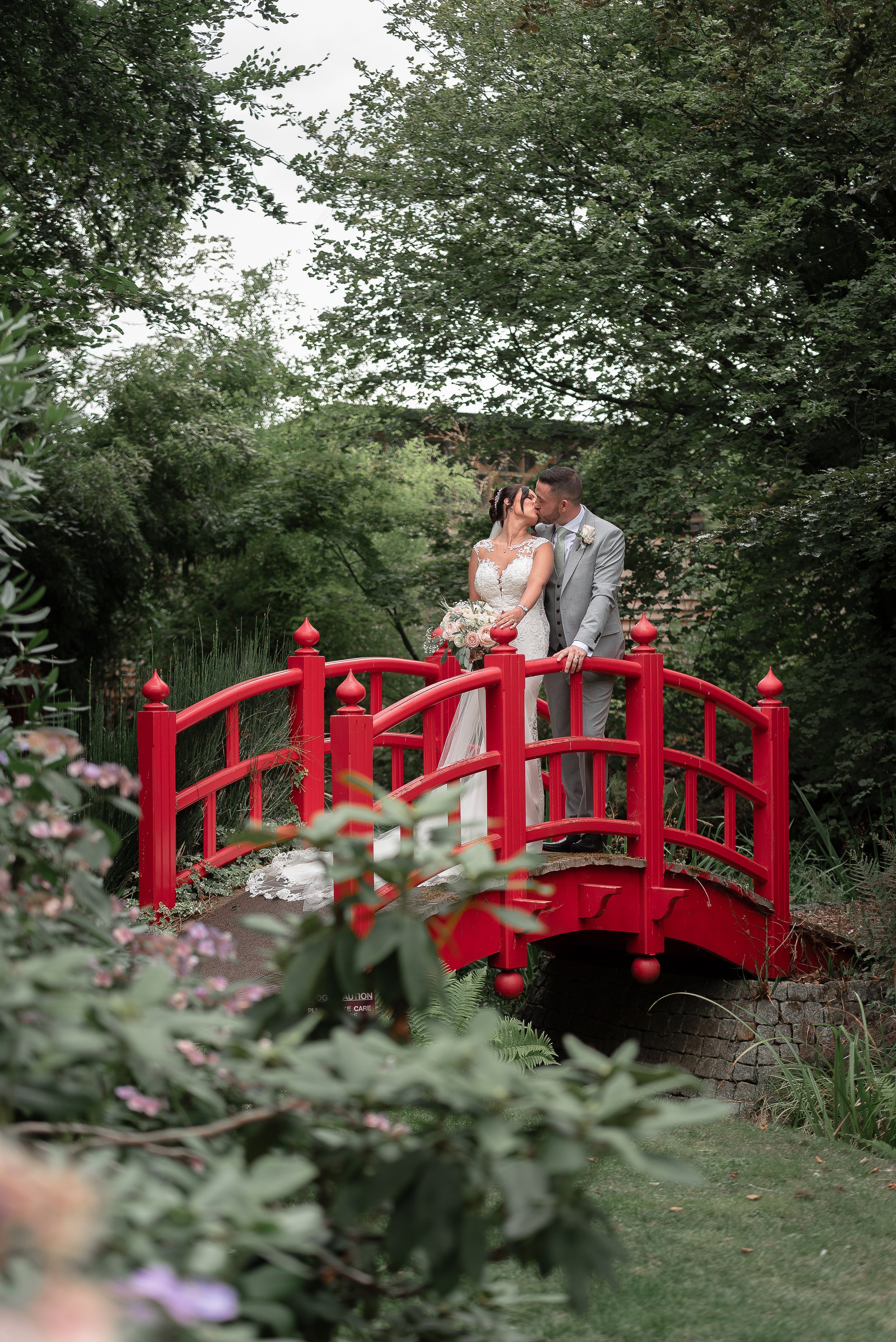 A bride and groom sharing a kiss on a small red bridge in a lush garden surrounded by greenery.