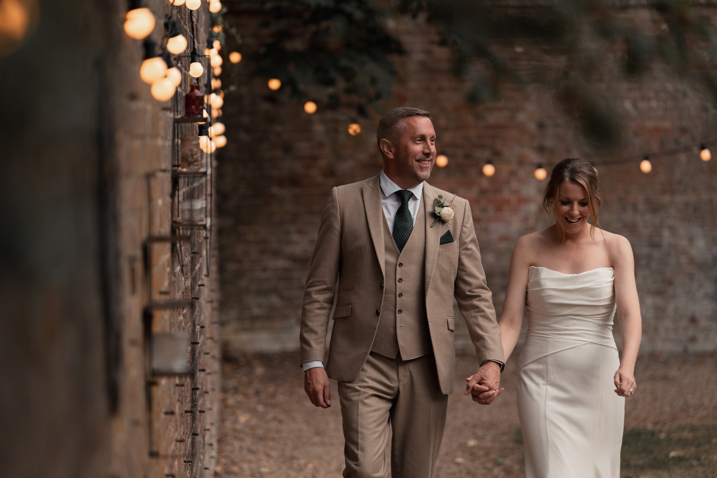 A happy couple, a man in a beige suit and a woman in a strapless white wedding dress, holding hands and smiling while walking outdoors near a brick wall with string lights.