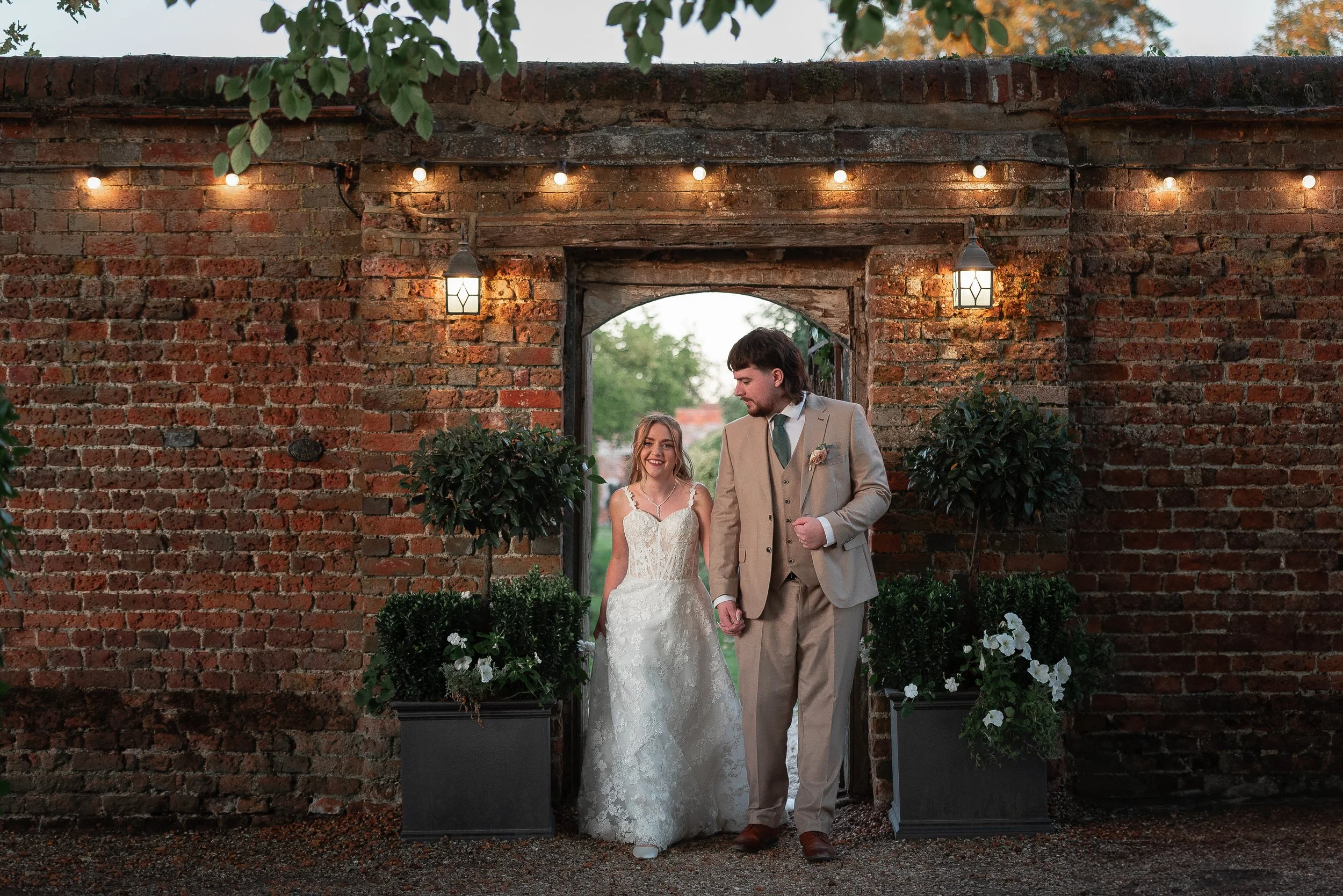 A bride and groom holding hands walking in front of a rustic brick archway decorated with white flowers and greenery, illuminated by warm string lights and lanterns during an outdoor wedding ceremony.