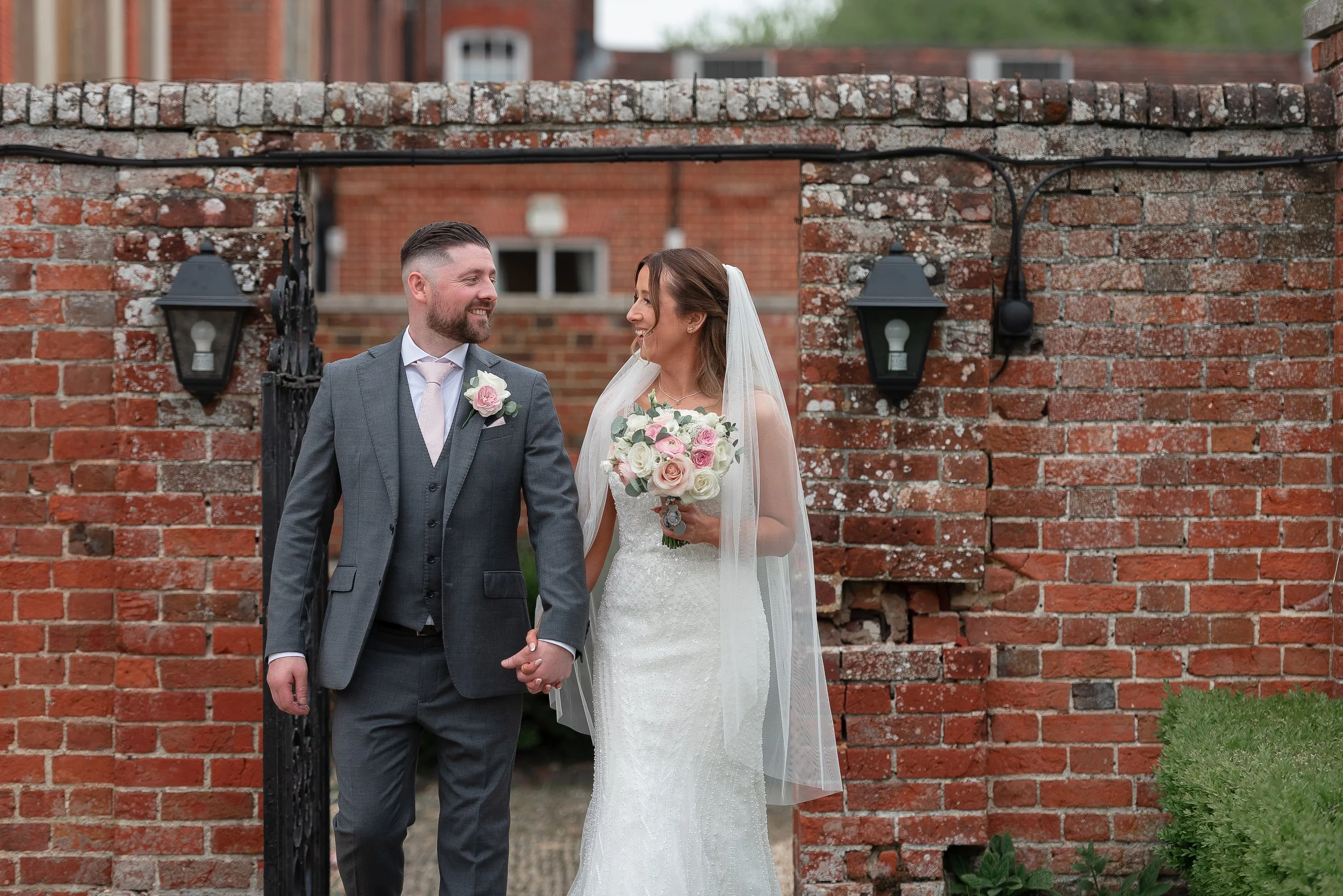 A bride and groom holding hands, smiling at each other, standing in front of a brick wall with black lantern-style lights, during their wedding ceremony.