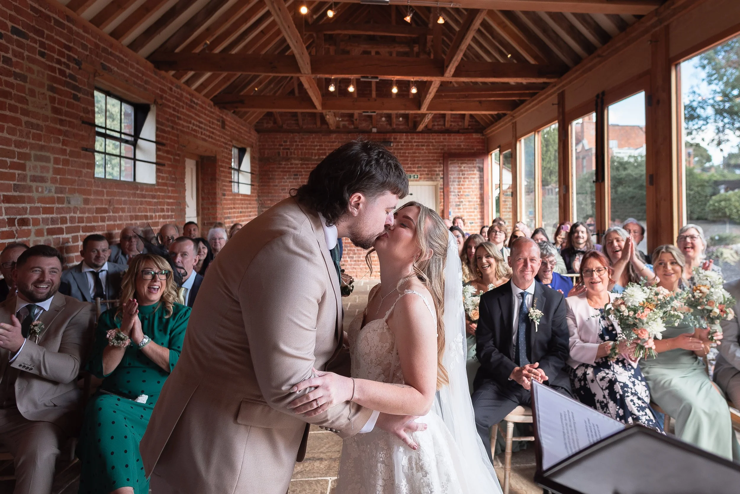 A couple is kissing during a wedding ceremony inside a rustic venue with brick walls and large windows. Guests are seated and clapping, smiling and enjoying the moment.