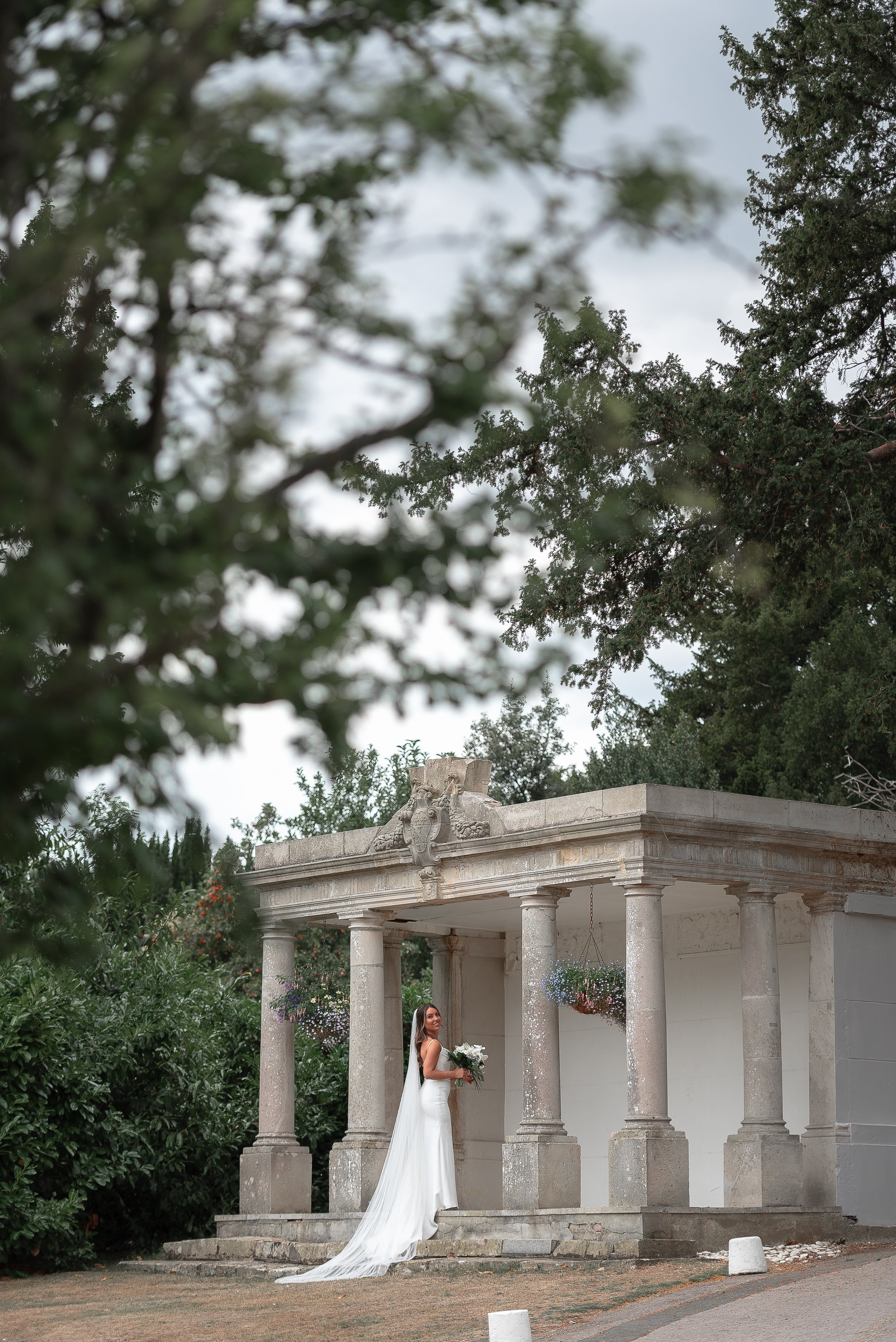 A bride in a white wedding dress holding a bouquet of flowers standing on the steps of a classical style building with columns, surrounded by lush greenery and trees.