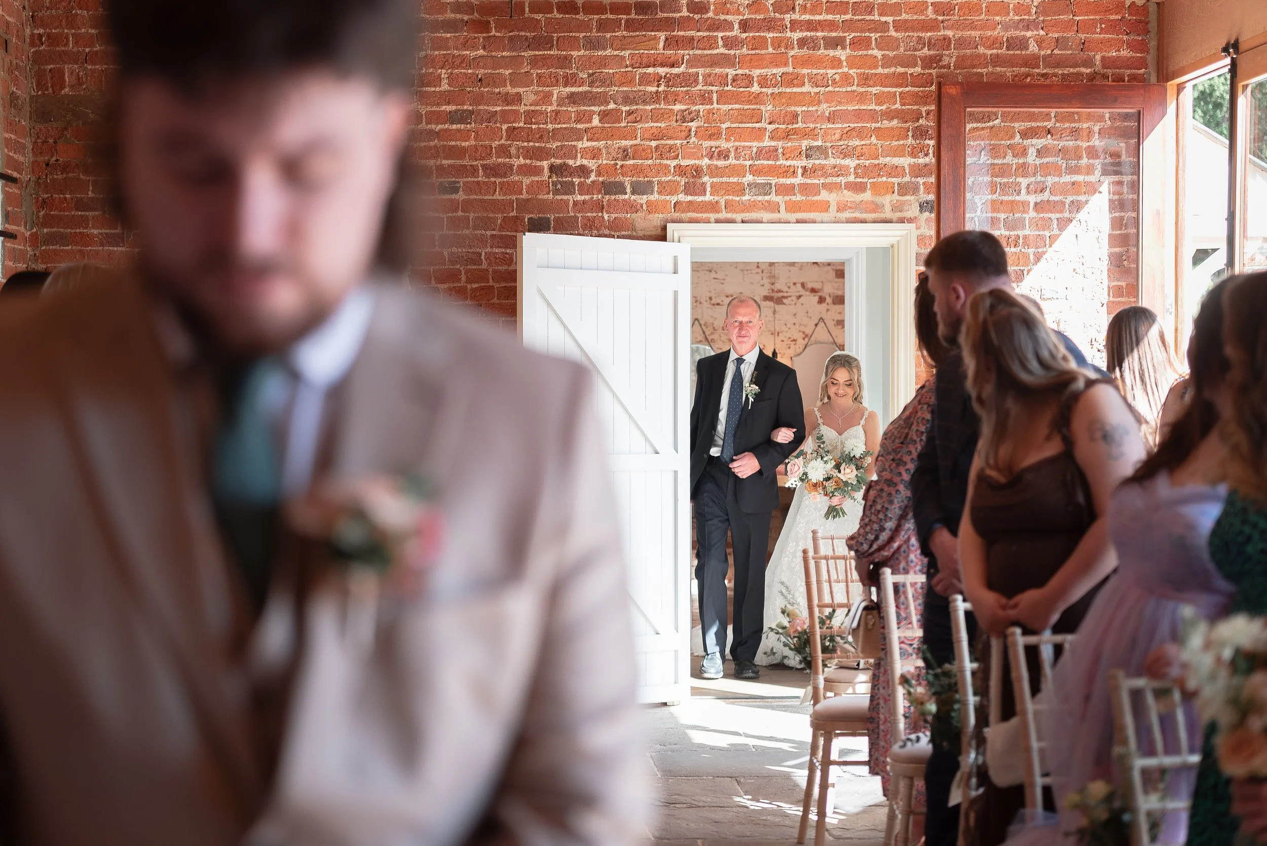 Wedding ceremony inside a brick building with guests seated on wooden chairs, the bride and groom are entering through a white door, with the groom escorting the bride.