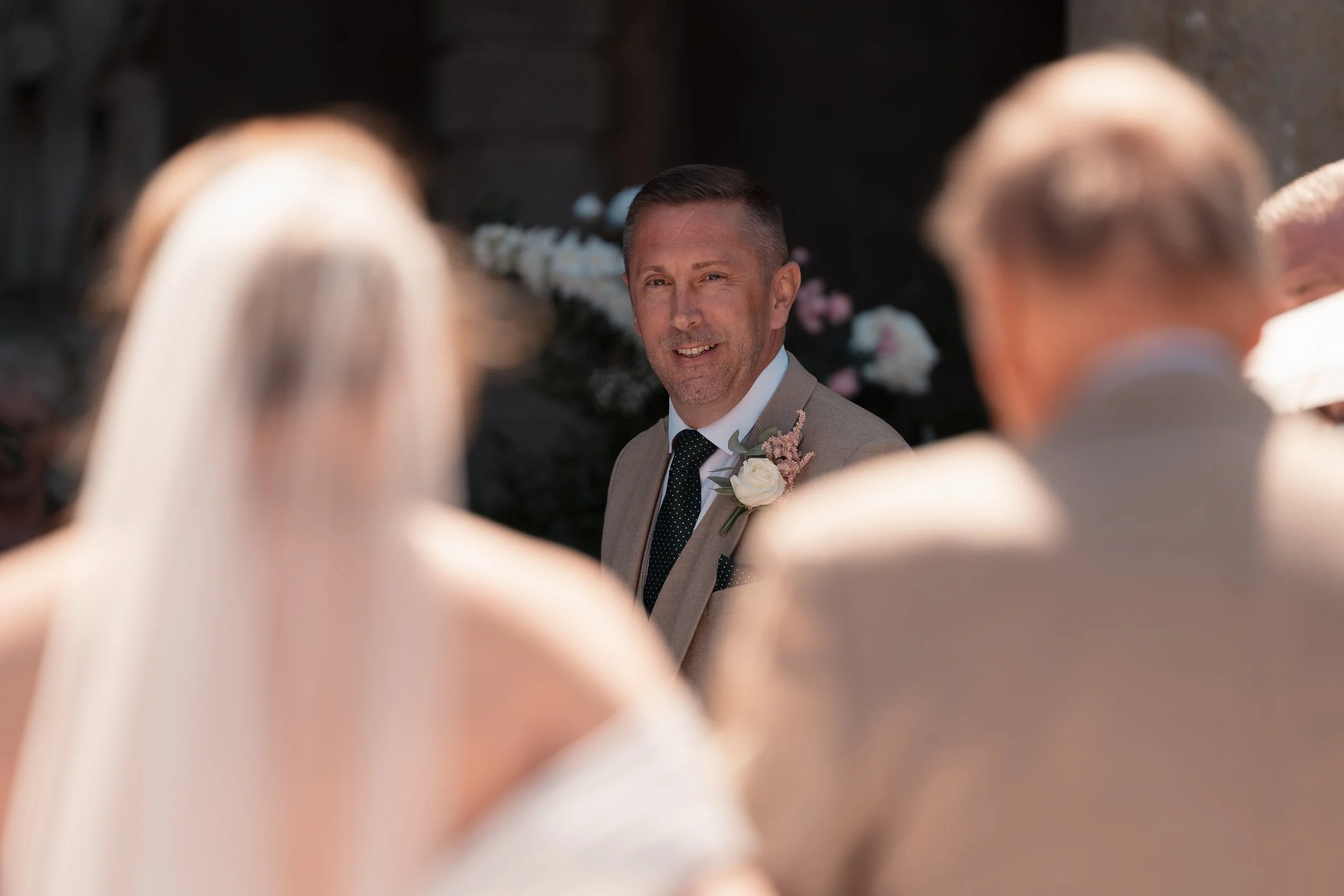 A man in a suit with a boutonniere looks at people in white dresses, possibly at a wedding ceremony.