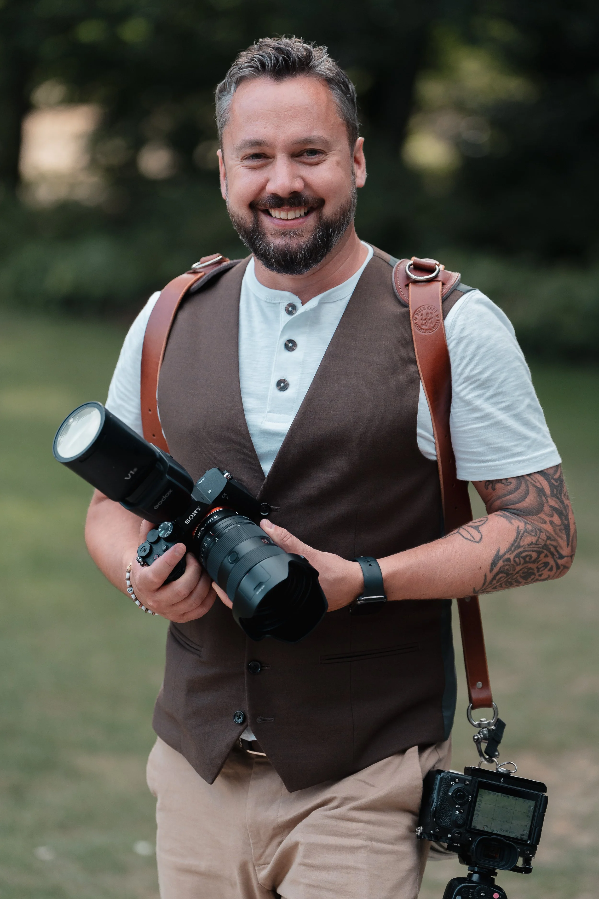 Paul Stickland, wearing a white shirt and brown vest, holding a camera with a large lens, outdoors in a park, smiling at the camera.
