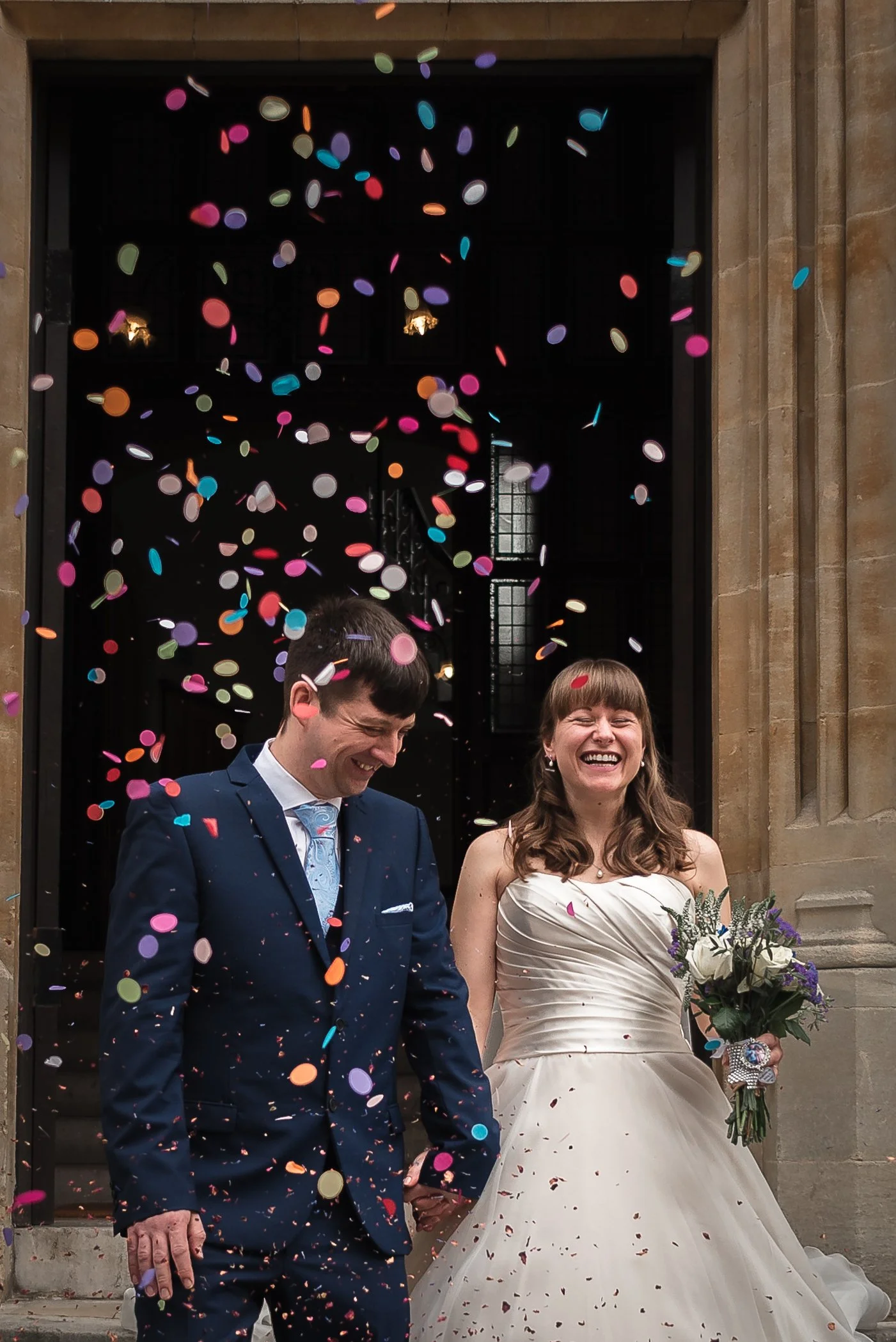 A bride and groom celebrating their wedding outside Winchester Registry Office with colourful confetti falling around them, smiling and holding hands.