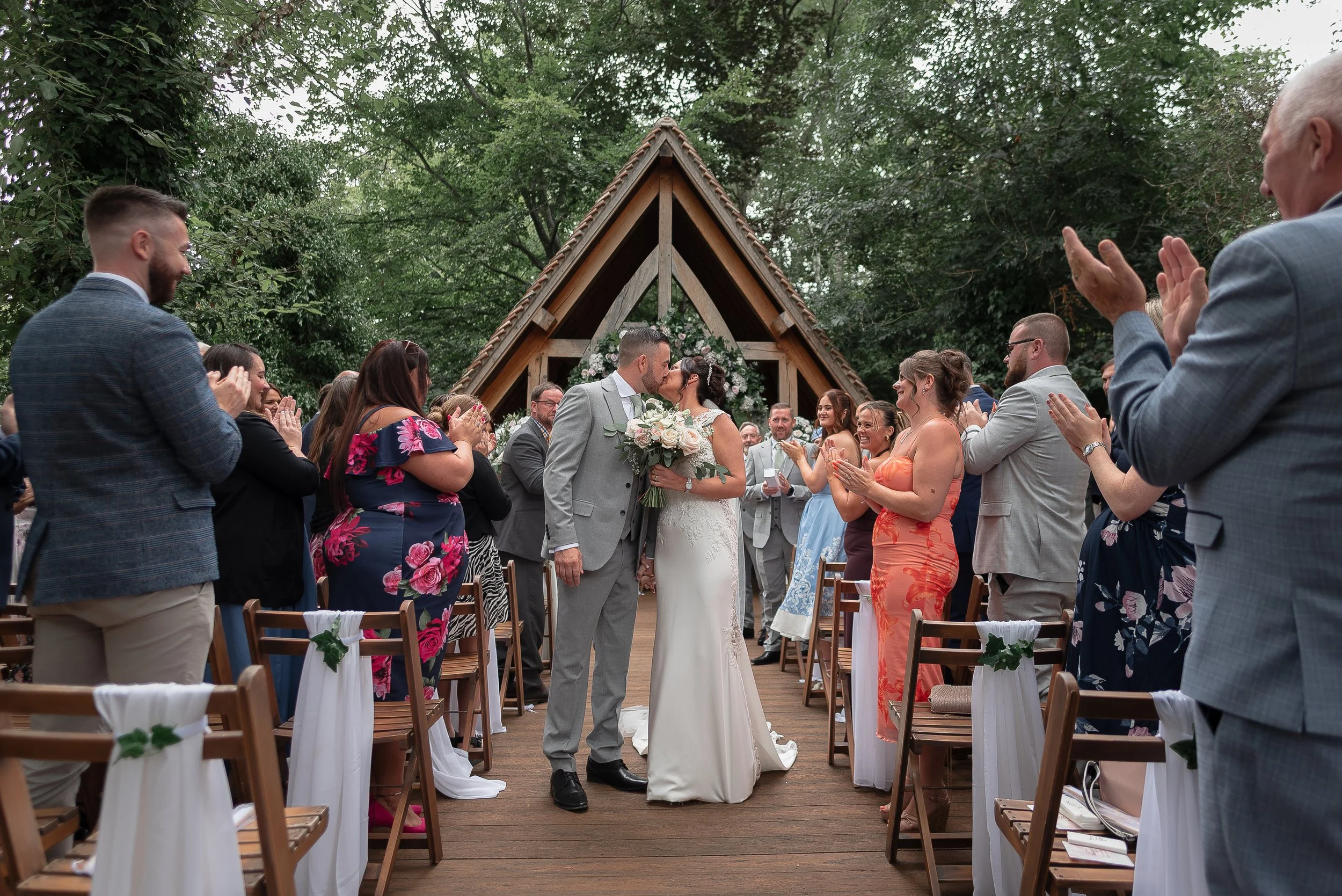 A bride and groom kiss at an outdoor wedding ceremony with friends and family clapping and smiling around them. The ceremony takes place under a wooden A-frame structure decorated with flowers, surrounded by green trees.