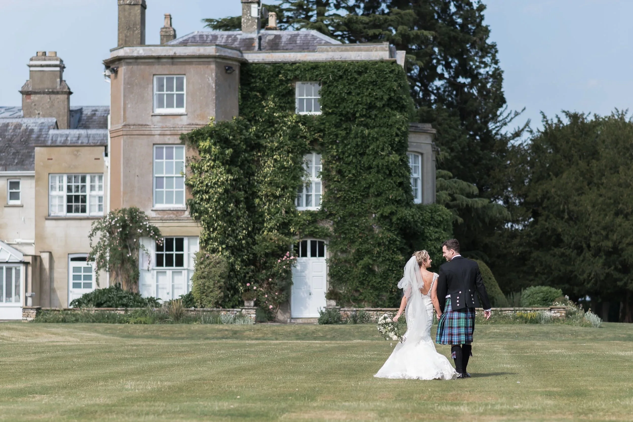 just married, the bride and groom walking hand in hand in the garden of Northbrook Park after just getting married. Groom is wearing a Scottish Kilt and the bride in a stunning white dress holding her bouquet.
