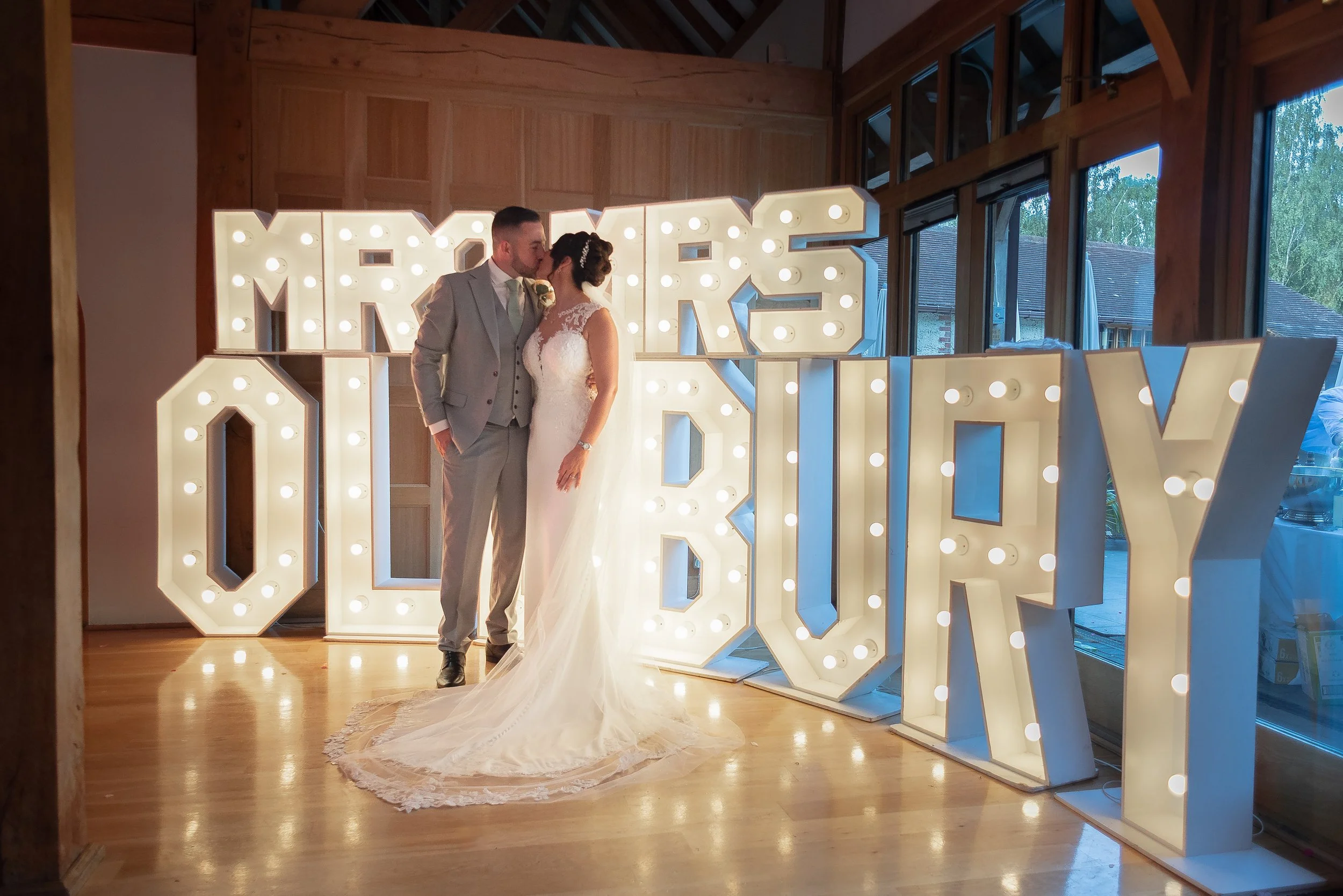 A bride and groom sharing a kiss in front of illuminated large decorative letters that spell their surname at a Hampshire wedding reception, The Rivervale Barn,  inside a wooden venue with large windows and natural light.