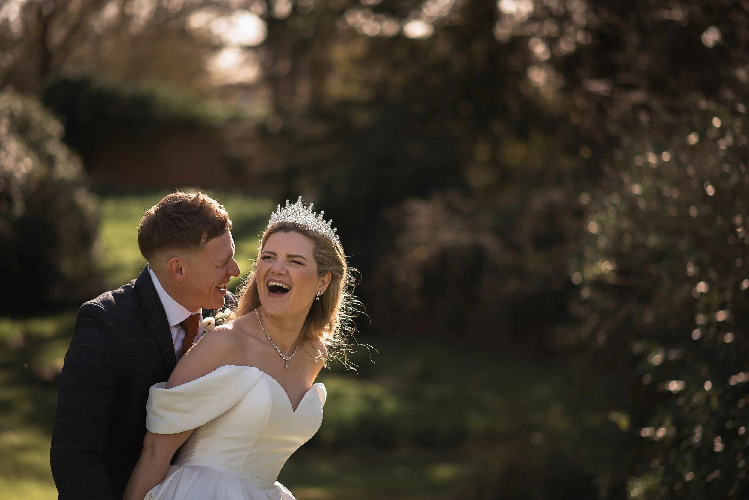 A happy newly wed couple outdoors on their wedding day, with the woman wearing a tiara and white dress, and the man in a suit, smiling and laughing during golden hour