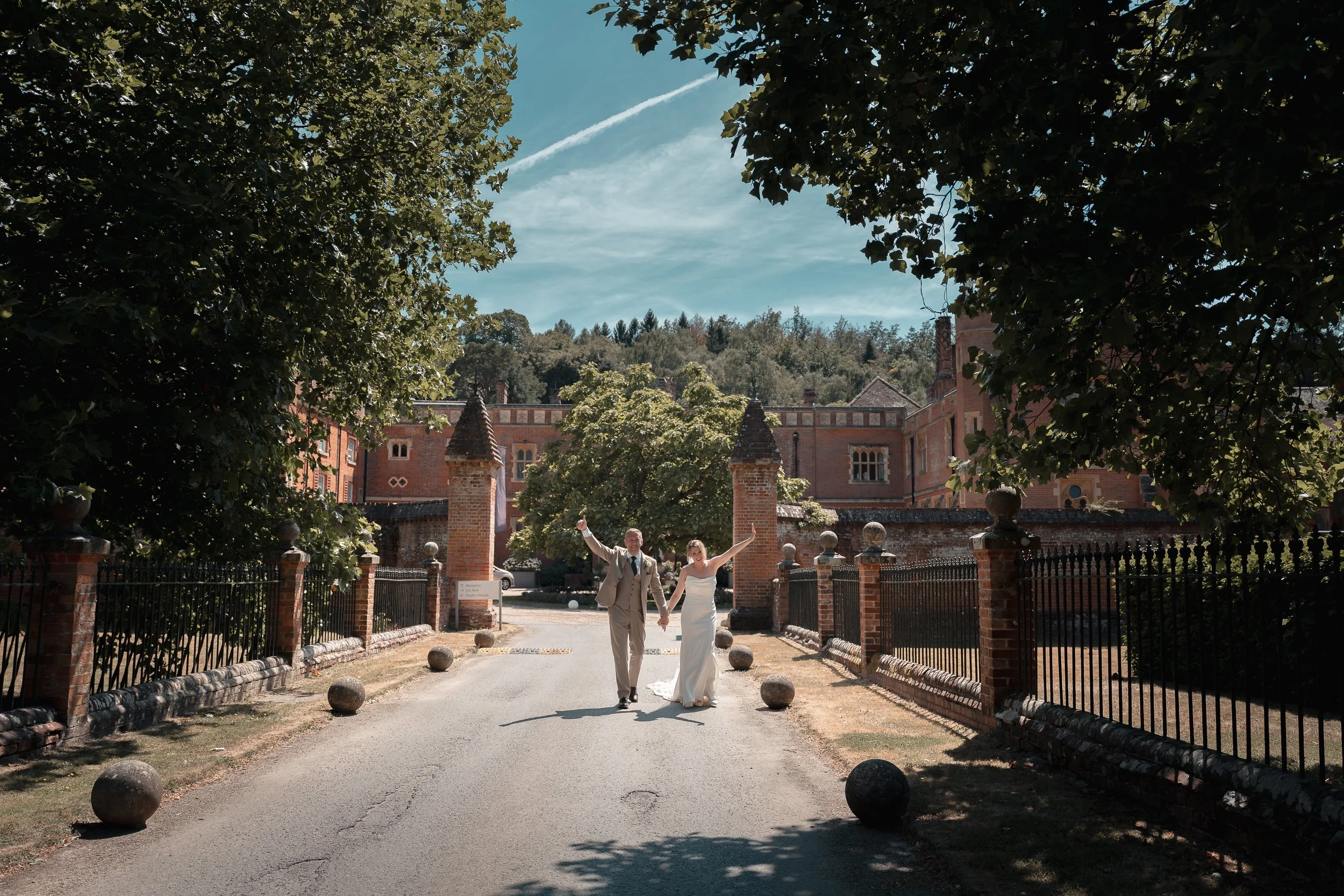 A bride and groom walking hand in hand outside a large red brick castle building, surrounded by trees and a fence, with a clear blue sky and some clouds overhead.