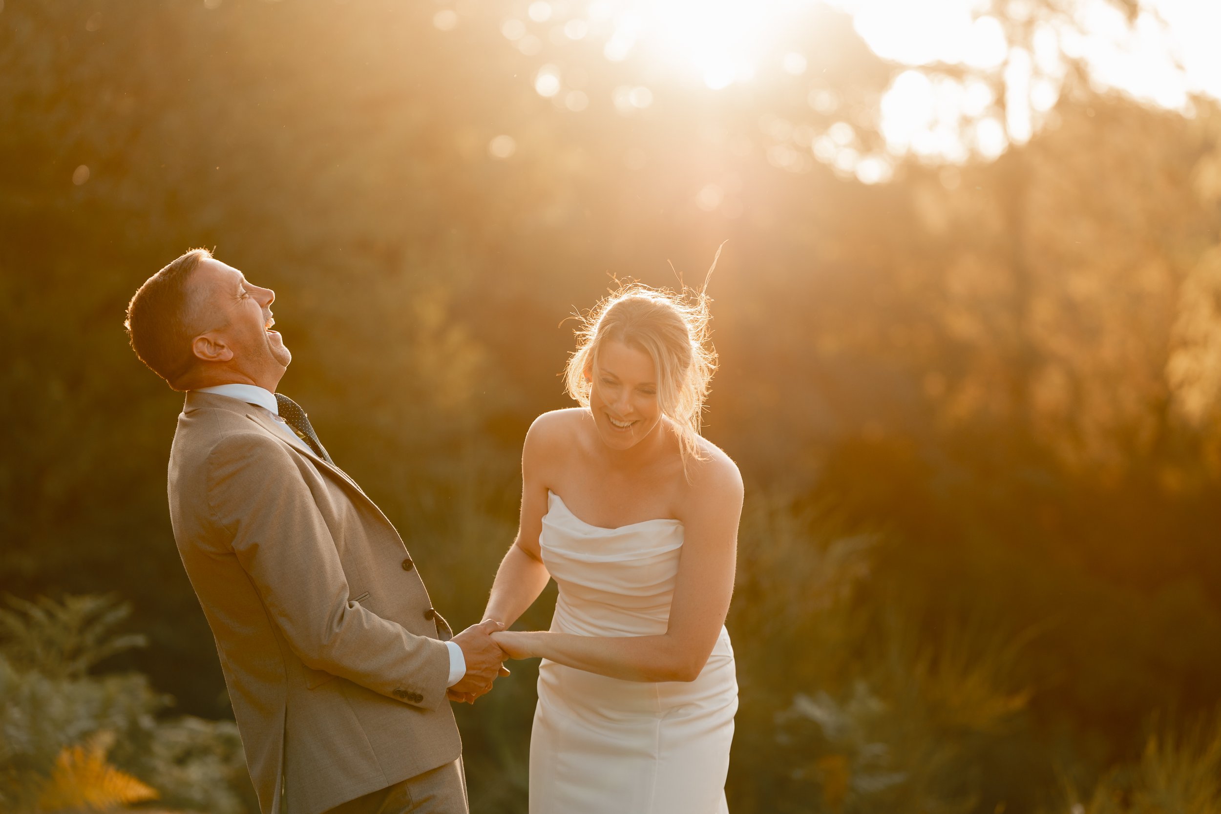 A man and a woman are smiling and holding hands outdoors during sunset, with the woman wearing a strapless white dress and the man in a beige suit.